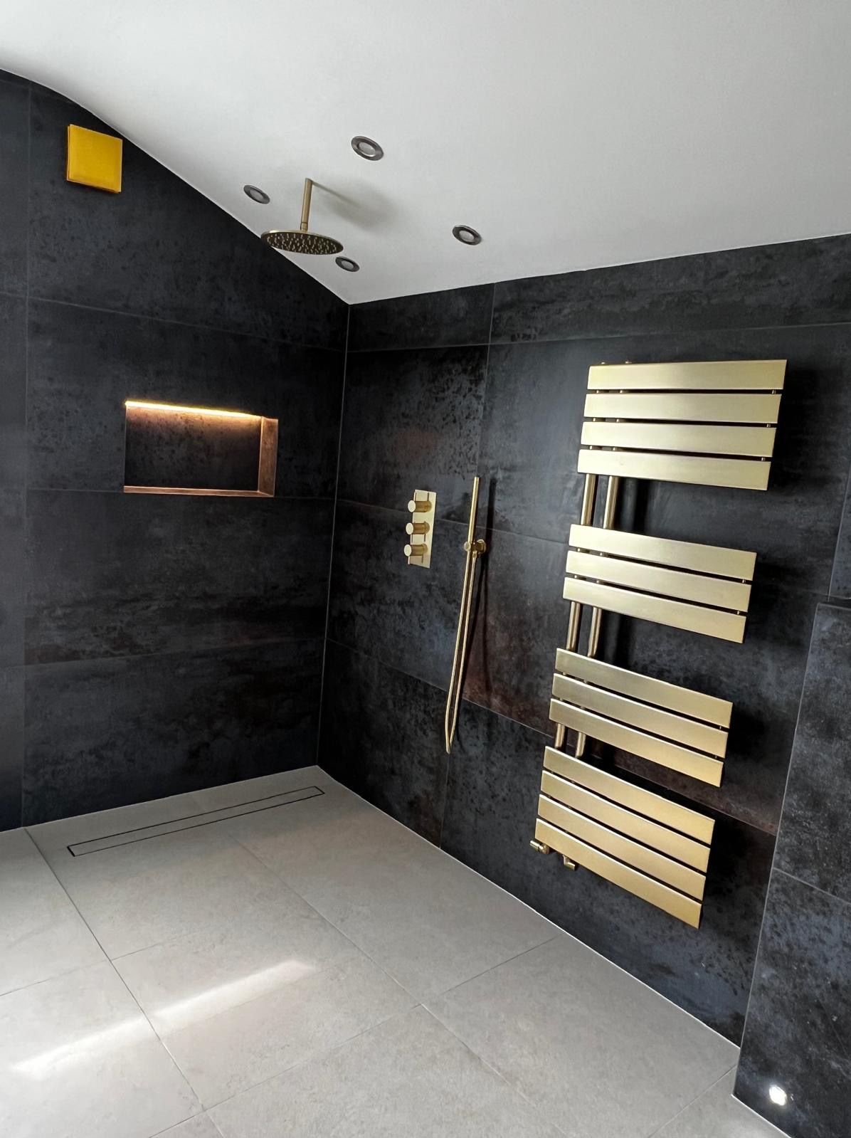 Nowoczesna łazienka z ciemnymi płytkami, złotą armaturą, grzejnikiem drabinkowym i wnęką z podświetleniem LED.