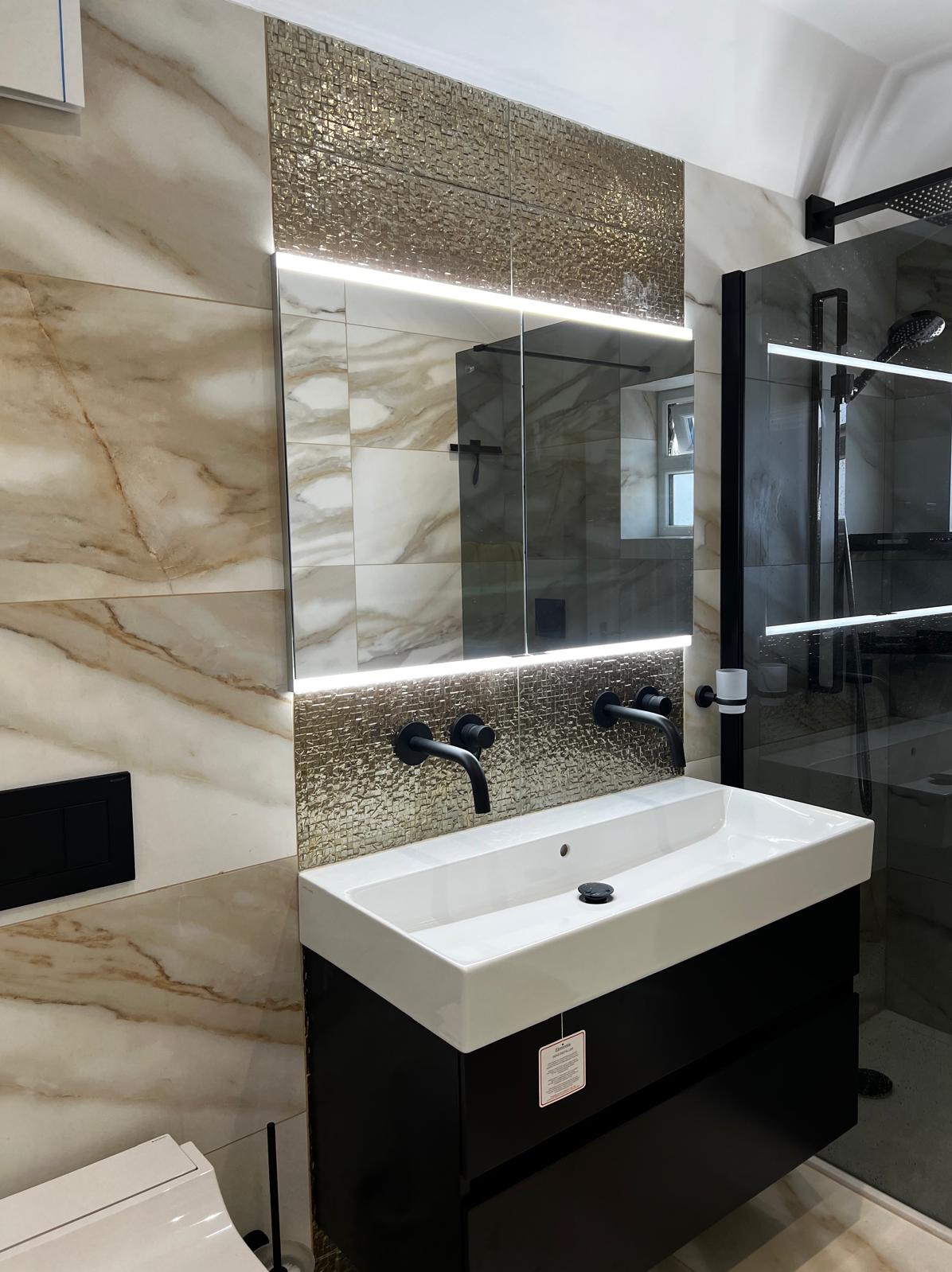 Nowoczesna łazienka z umywalką nablatową na czarnej szafce, czarne baterie, lustro z podświetleniem LED i mozaiką w kolorze złota oraz kabina prysznicowa z czarnymi profilami.