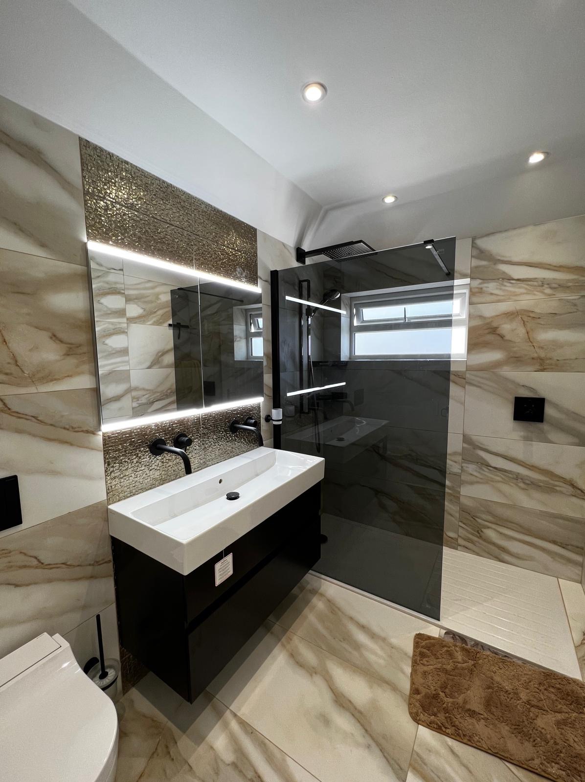 Nowoczesna łazienka z kabiną prysznicową typu walk-in, umywalką nablatową na czarnej szafce, lustrem z podświetleniem LED i płytkami imitującymi marmur.