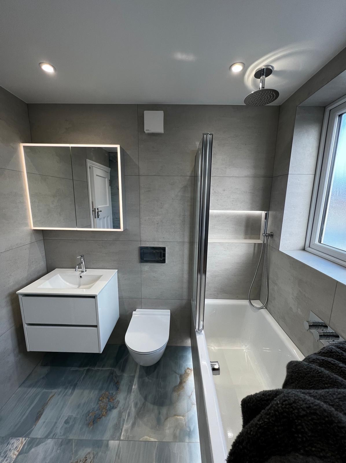 Nowoczesna łazienka z szarymi płytkami, podświetlanym lustrem, białą szafką umywalkową, toaletą i wanną z parawanem, podkreślona niebiesko-złotą podłogą z płytek imitujących kamień.