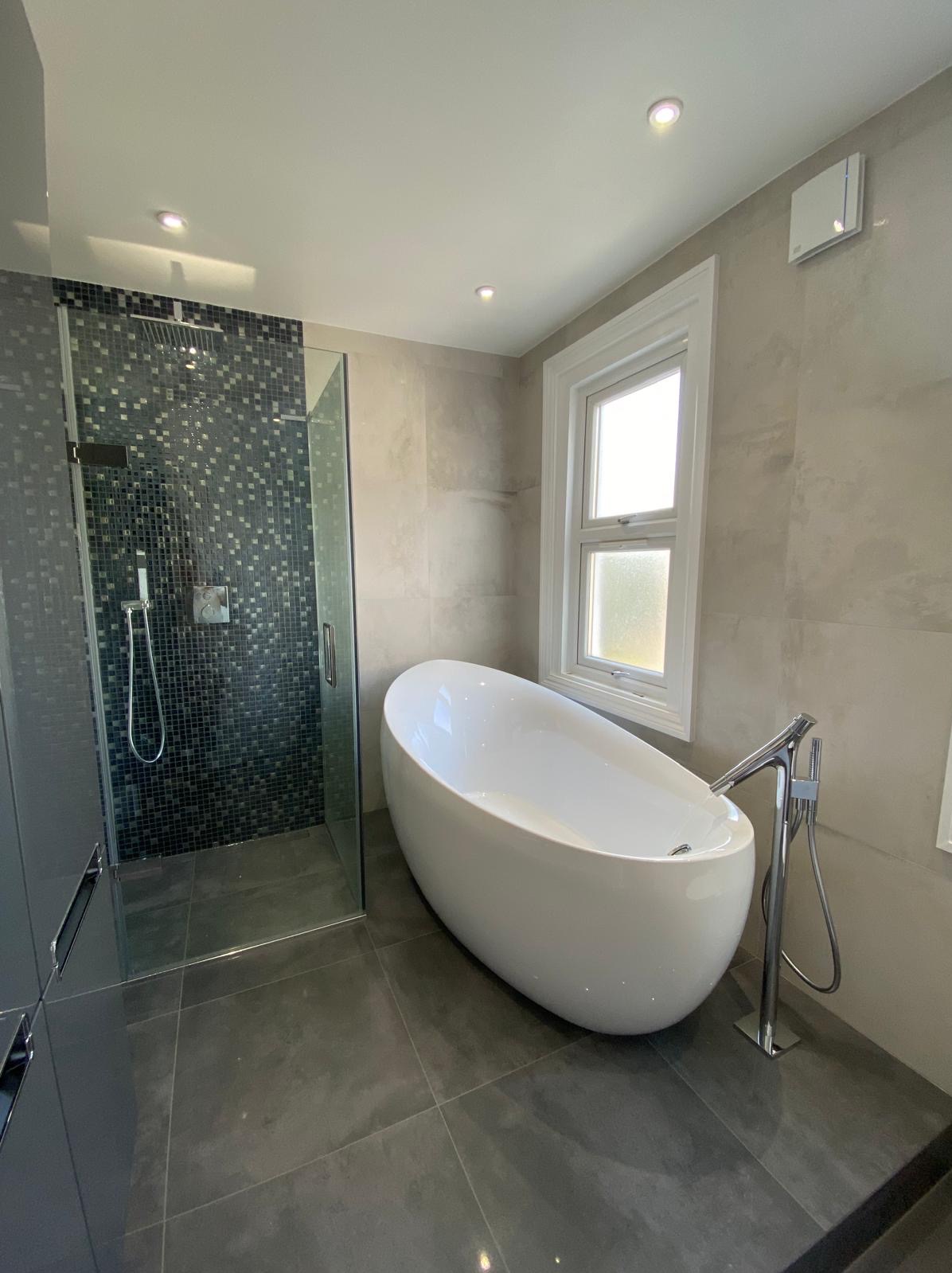 Nowoczesna łazienka z wolnostojącą białą wanną, kabiną prysznicową z mozaiką i szarą glazurą ścienną oraz podłogową, oświetlona punktowymi lampami sufitowymi.