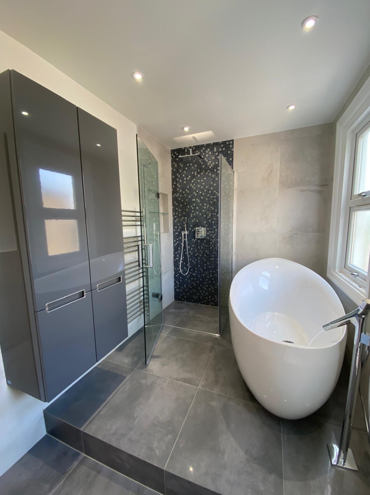 Nowoczesna łazienka z wolnostojącą wanną, kabiną prysznicową z mozaiką, szarą zabudową meblową na wysoki połysk i grzejnikiem drabinkowym