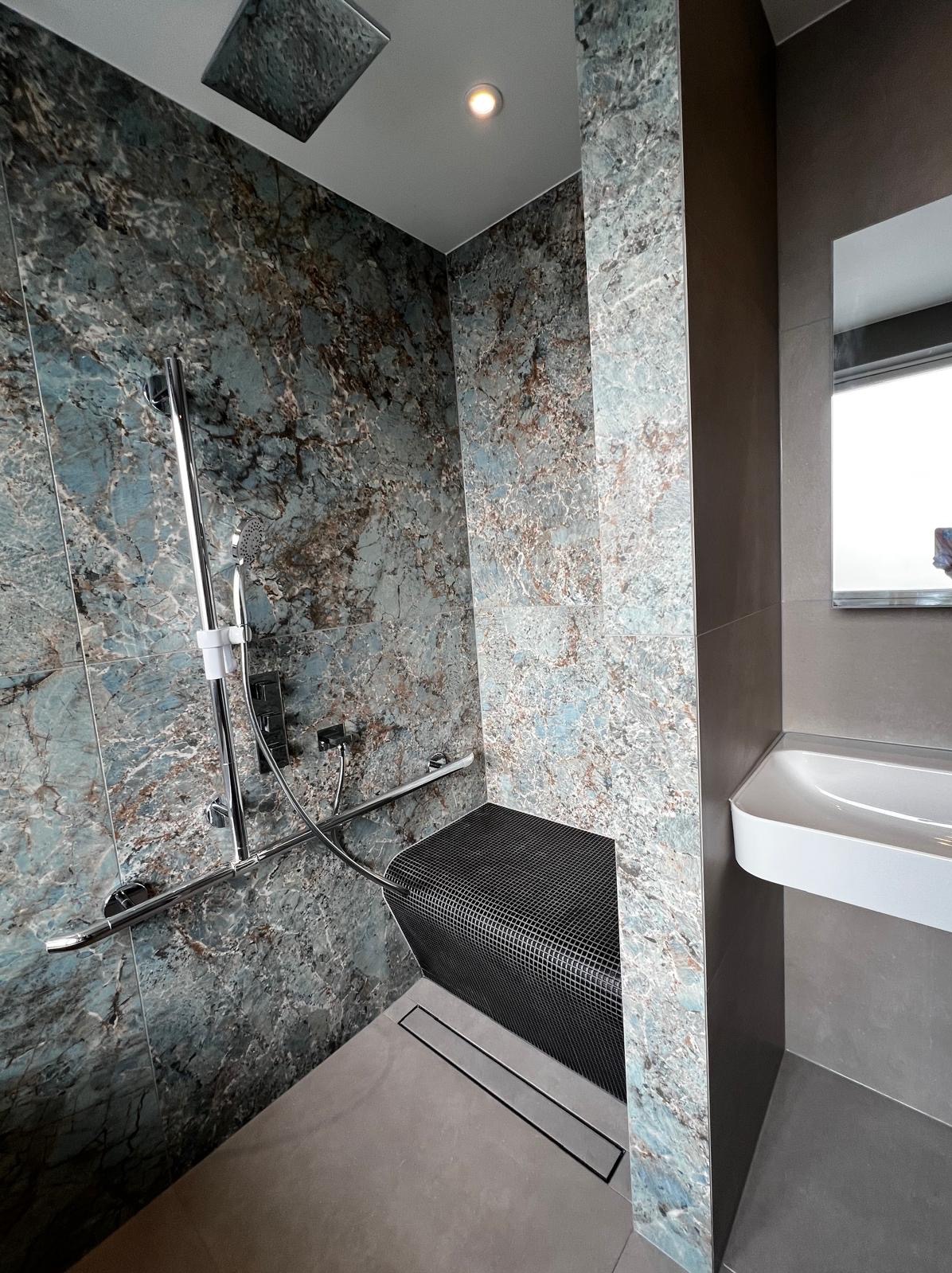 Nowoczesna kabina prysznicowa z siedziskiem wykończona płytkami imitującymi marmur, chromowaną armaturą i odpływem liniowym w posadzce, obok umywalka nablatowa.