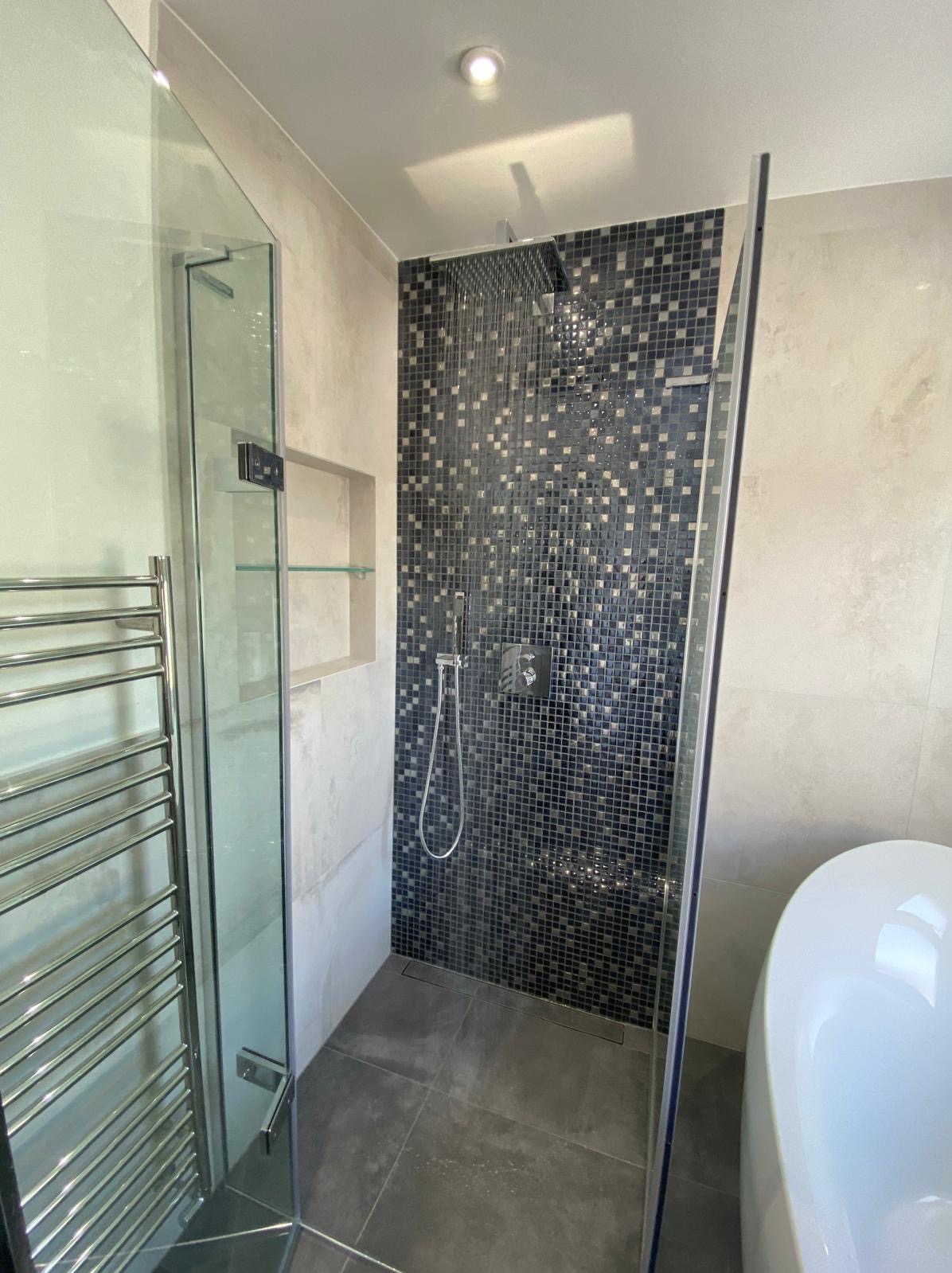 Nowoczesna kabina prysznicowa z mozaiką w odcieniach szarości i czerni, szklane drzwi, grzejnik drabinkowy i fragment białej wanny w tle.