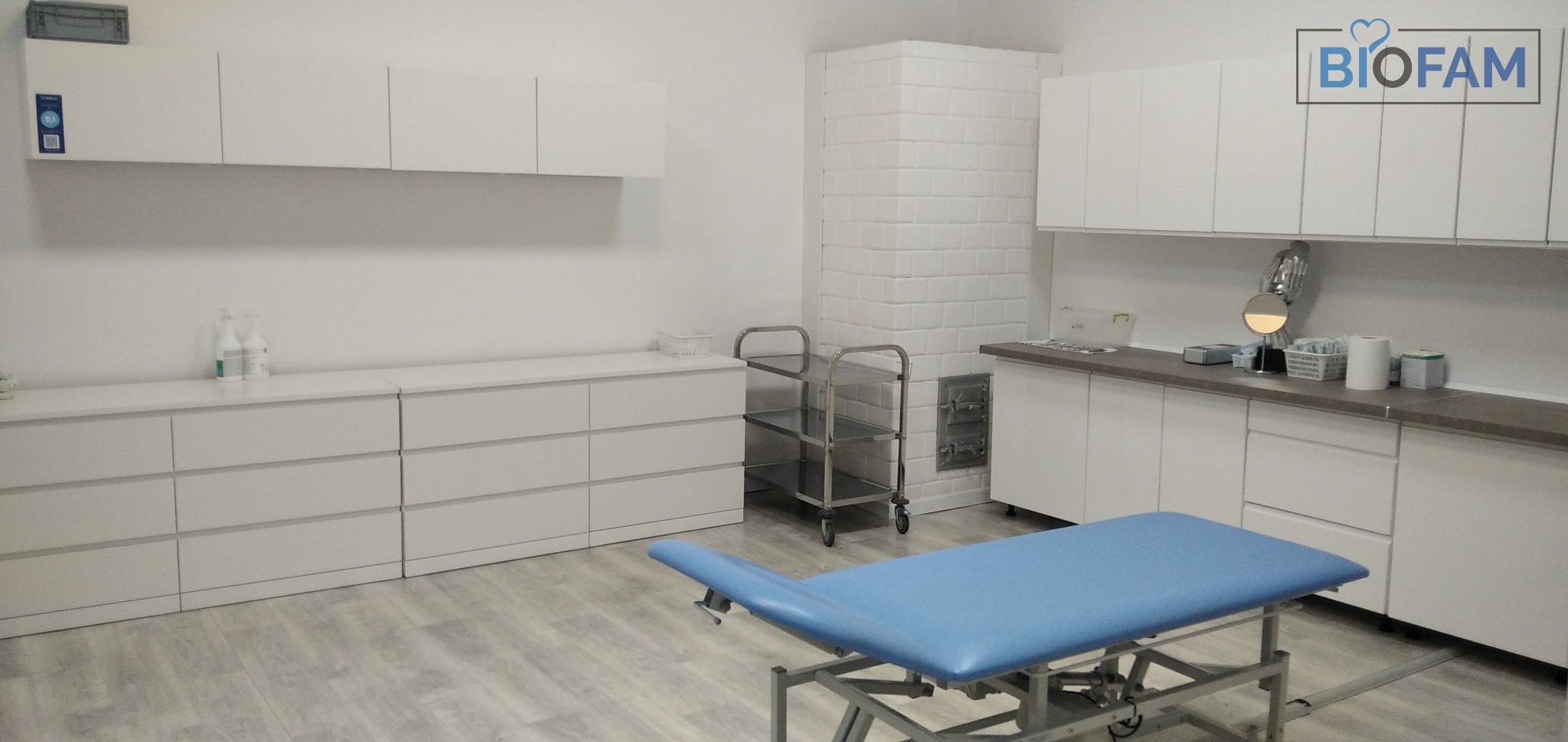 Jasne wnętrze gabinetu medycznego z błękitnym leżankiem, białymi szafkami, wózkiem na kółkach i piecem kaflowym w tle, logo Biofam w prawym górnym rogu.