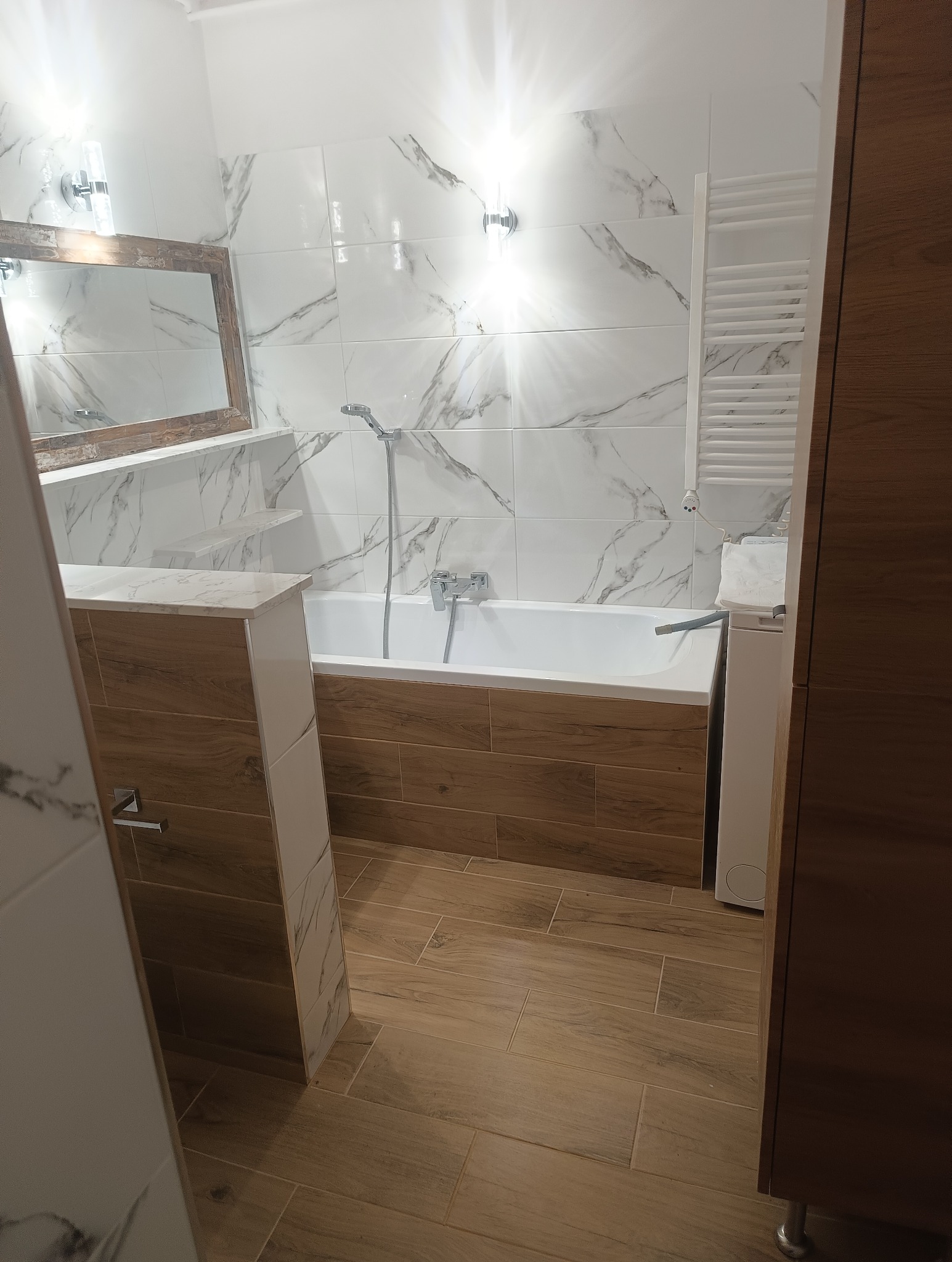 Wykończona łazienka w Piasecznie z wanną obudowaną drewnopodobnymi panelami, białymi płytkami imitującymi marmur na ścianach i drewnianą ramą lustra.