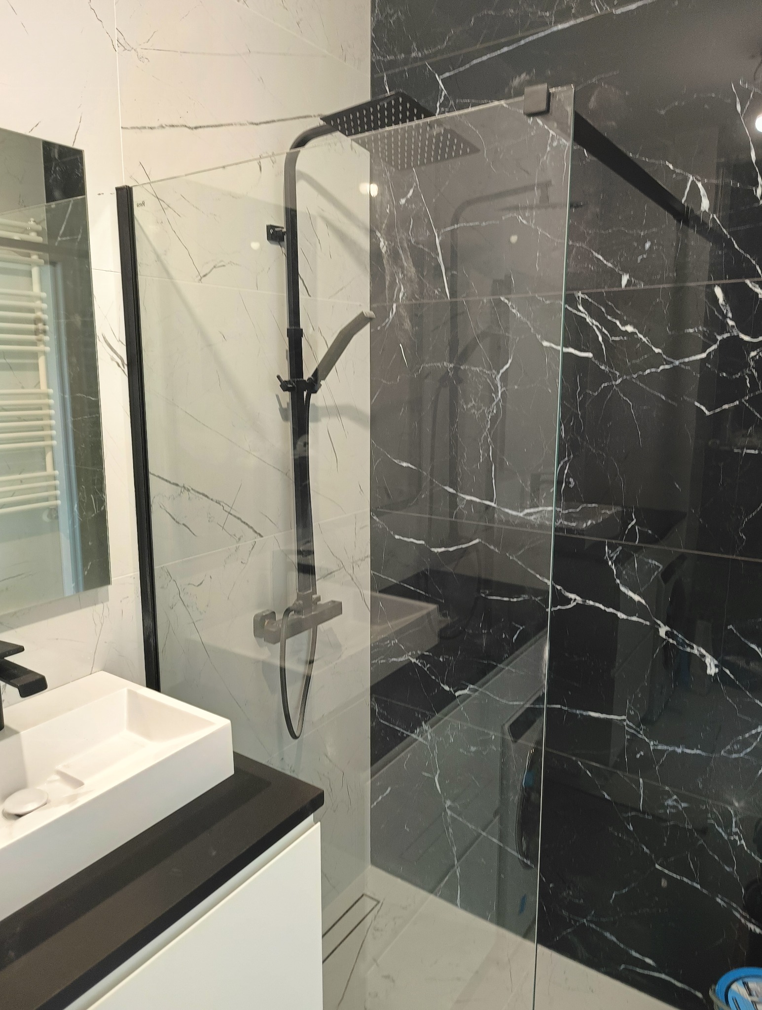 Nowoczesna łazienka z czarno-białymi marmurkowymi płytkami, kabiną prysznicową typu walk-in z czarną armaturą i białą umywalką nablatową na czarnym blacie.