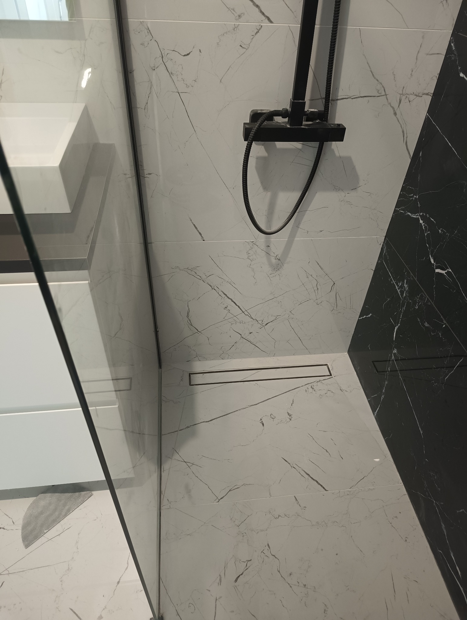 Nowoczesny prysznic walk-in z czarną armaturą, odpływem liniowym i płytkami imitującymi biały marmur z czarnymi żyłami, zestawiony z czarną płytką marmurową w rogu.