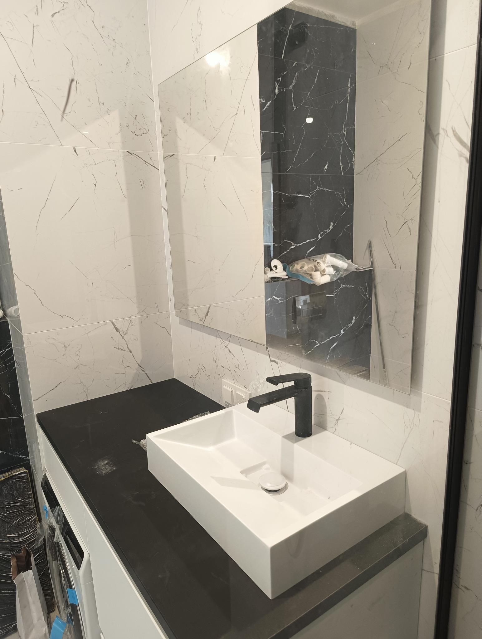 Nowoczesna łazienka w trakcie wykańczania: biała umywalka nablatowa z czarną baterią na czarnym blacie, marmurowe płytki na ścianach, odbicie akcesoriów w lustrze.