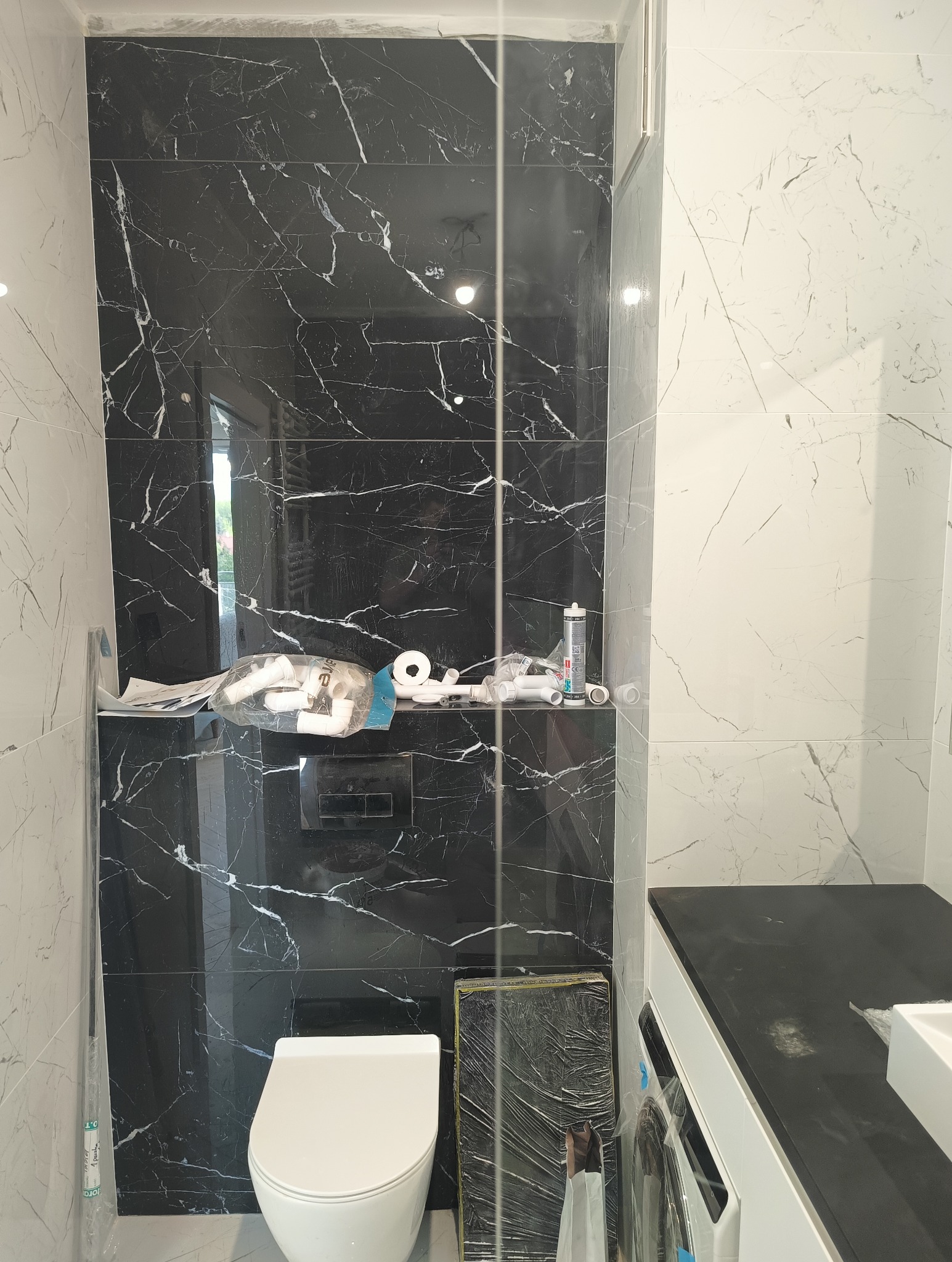 Częściowo wykończona łazienka z czarno-białymi marmurkowymi płytkami, wiszącą toaletą, oraz akcesoriami instalacyjnymi na półce.