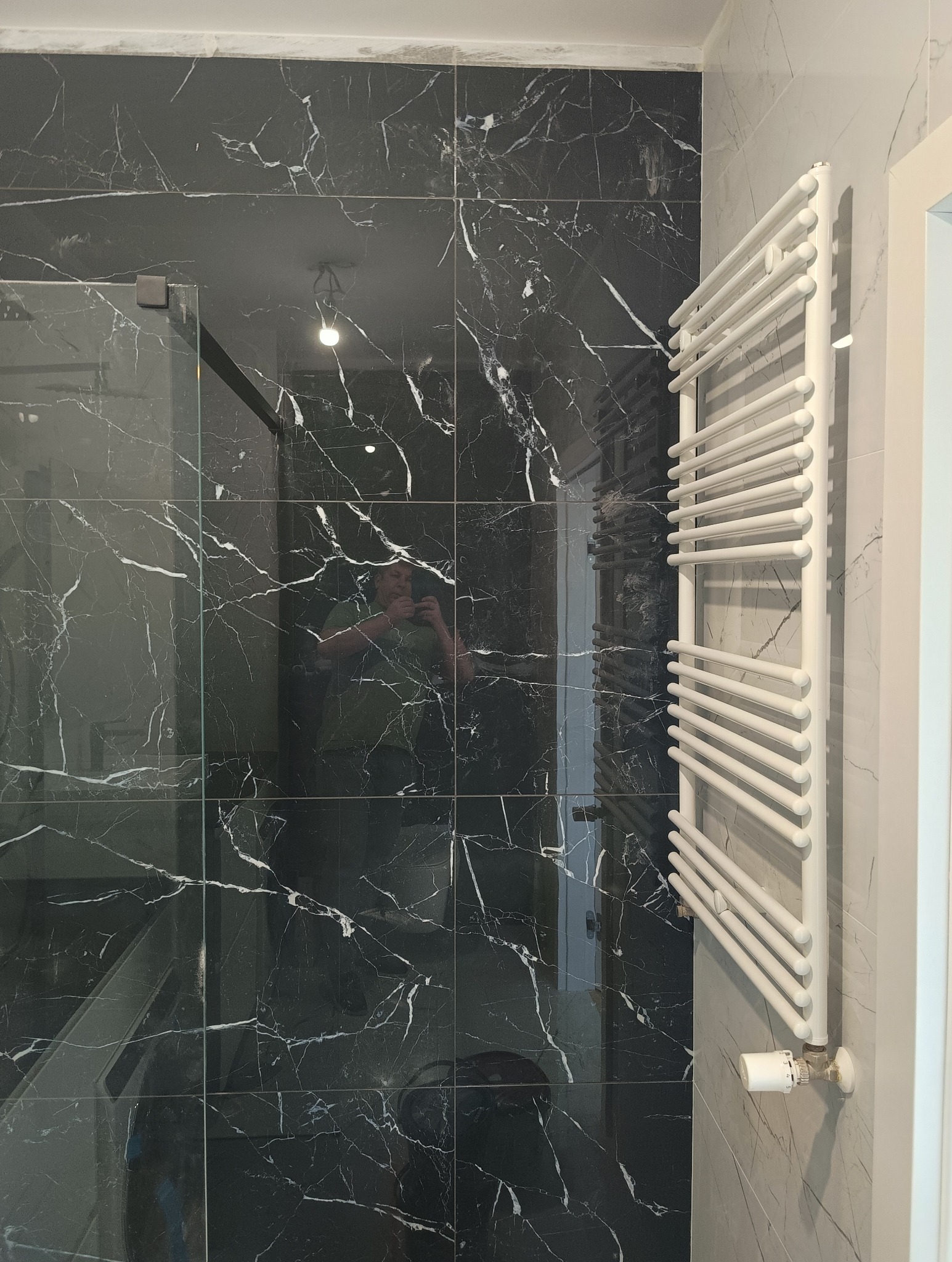Nowoczesna łazienka z czarnymi płytkami imitującymi marmur, kabiną prysznicową ze szklanymi drzwiami i białym grzejnikiem drabinkowym z termostatem.