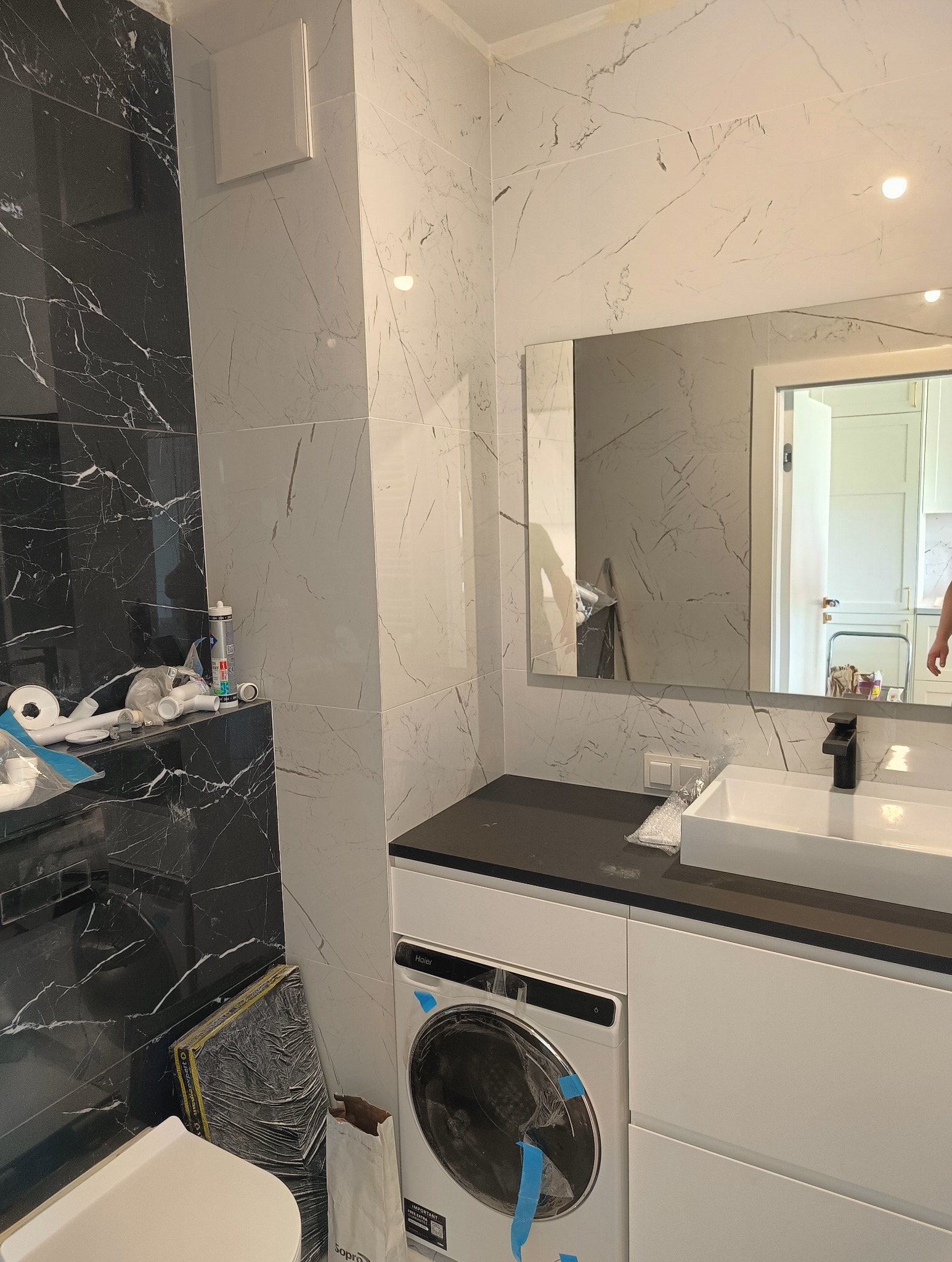 Łazienka w trakcie remontu: biało-czarne marmurowe płytki, biała umywalka z czarną armaturą, pralka Haier z niebieską taśmą zabezpieczającą, widoczne narzędzia i materiały budowlane.