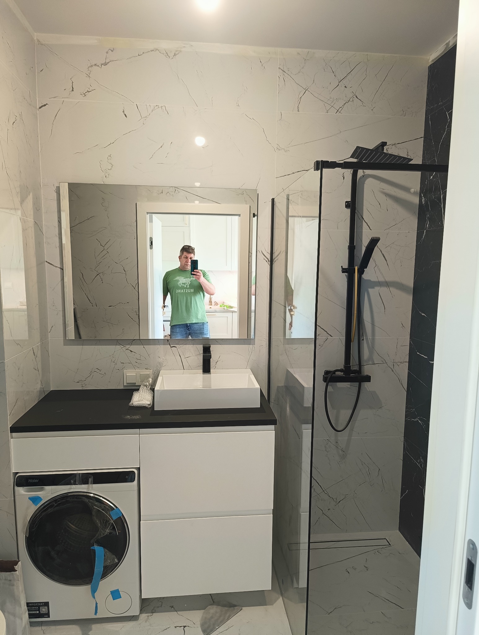 Nowoczesna łazienka z marmurowymi płytkami, kabiną prysznicową typu walk-in z czarną armaturą, umywalką nablatową na szafce z pralką Haier, widok z lustra na osobę robiącą zdjęcie w kuchni.