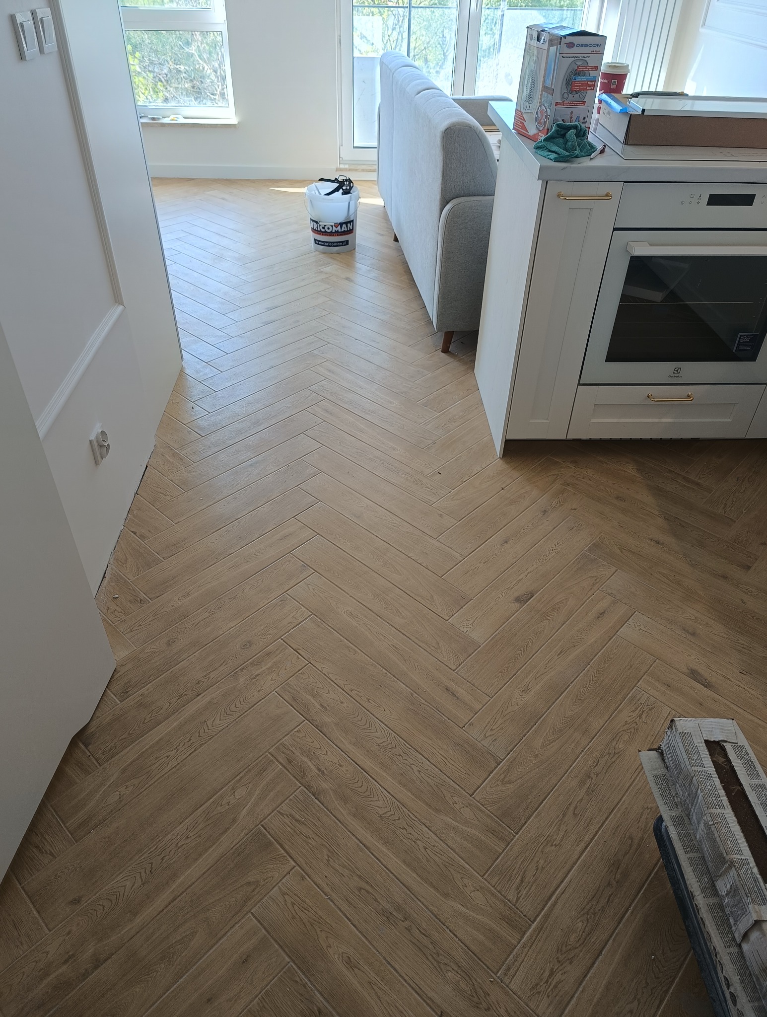 Ułożona podłoga z płytek imitujących drewno w jodełkę francuską, widoczny fragment kuchni i salonu, wiadro po kleju do płytek, świeżo po zakończeniu prac.