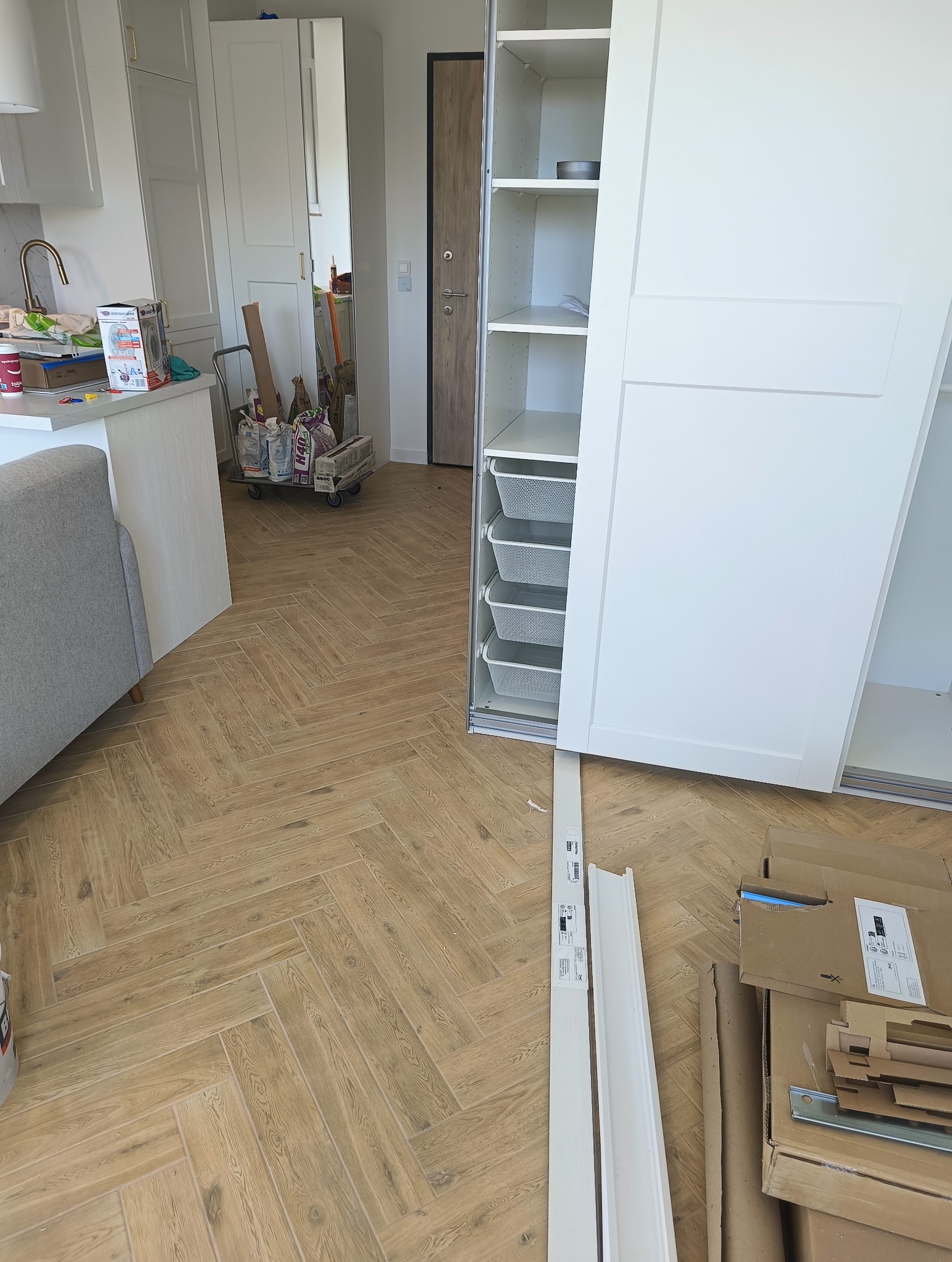 Montaż szafy wnękowej z białymi przesuwnymi drzwiami i metalowymi koszami wewnątrz, widoczne kartony po meblach i listwy przypodłogowe na podłodze z drewnopodobnych płytek ułożonych w jodełkę.