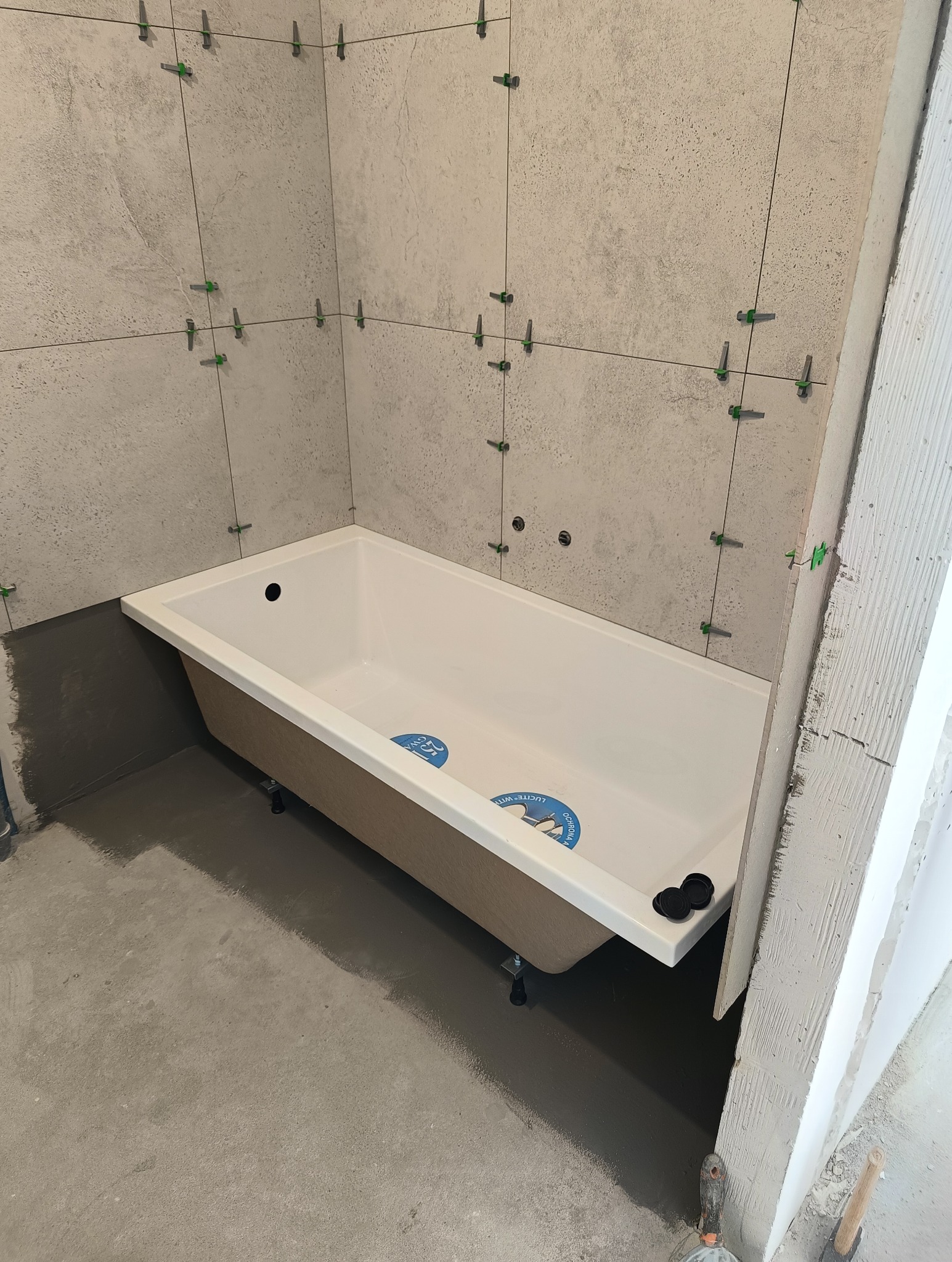 Wanna w trakcie montażu w surowej łazience z szarymi płytkami i systemem poziomowania, widoczne ślady kleju i narzędzia na podłodze.