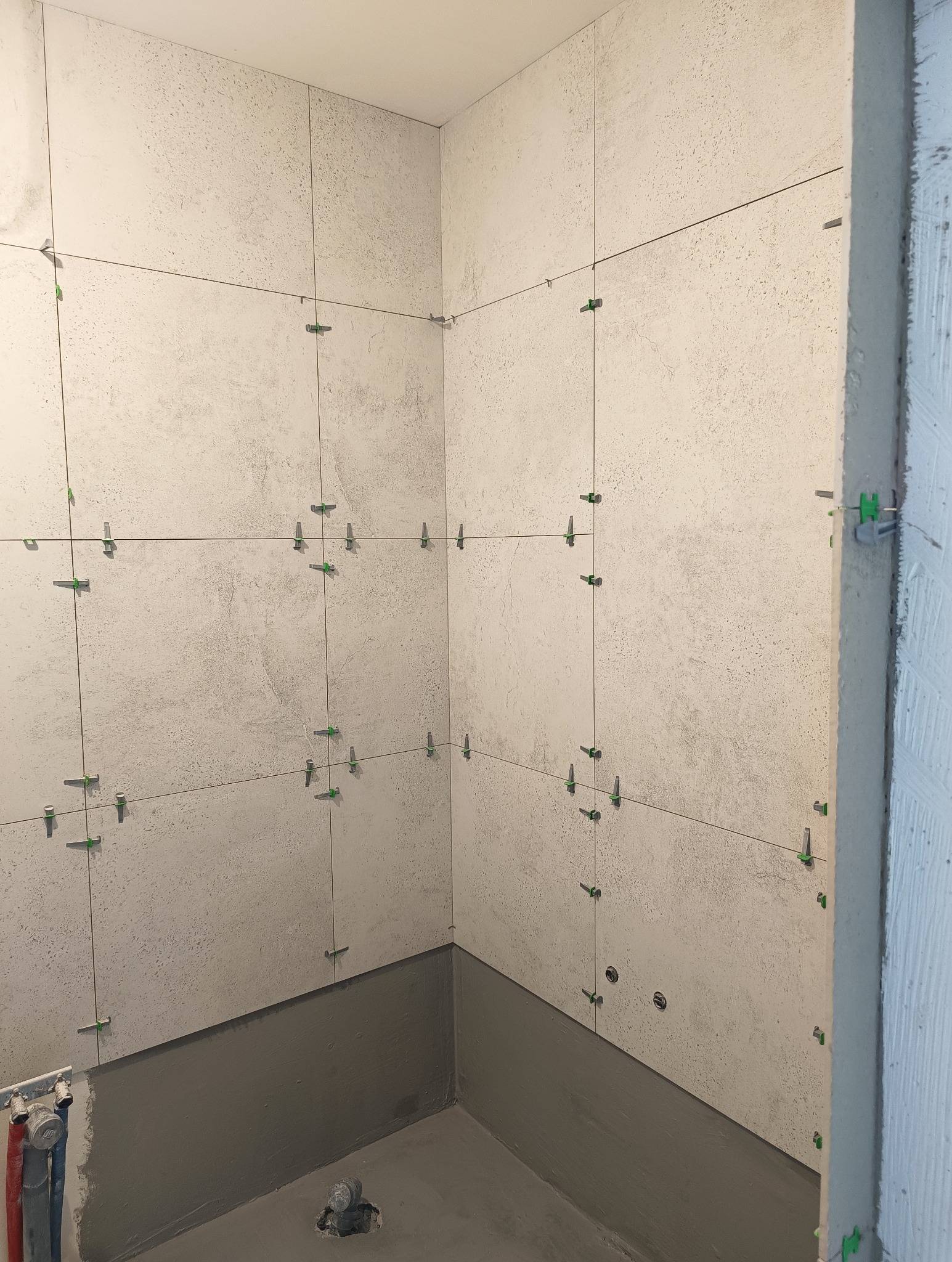 Świeżo ułożone, jasne płytki imitujące kamień na ścianach łazienki, z użyciem systemu poziomowania płytek (zielone klipsy i kliny), widoczne przygotowane otwory na instalacje i hydroizolacja przy...