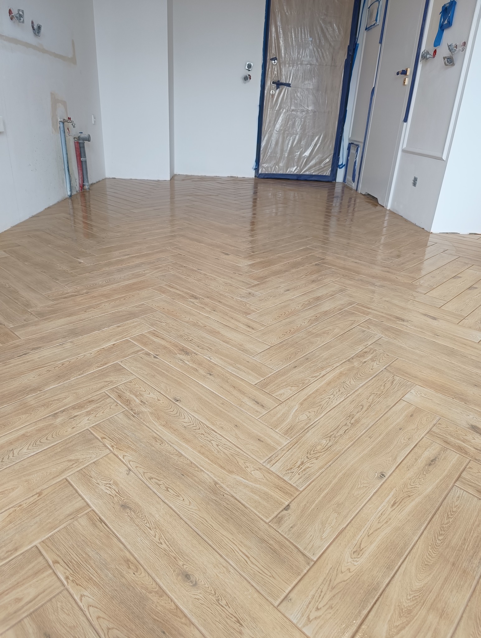 Ułożona podłoga z płytek imitujących drewno w jodełkę w trakcie remontu łazienki, drzwi zabezpieczone folią malarską.