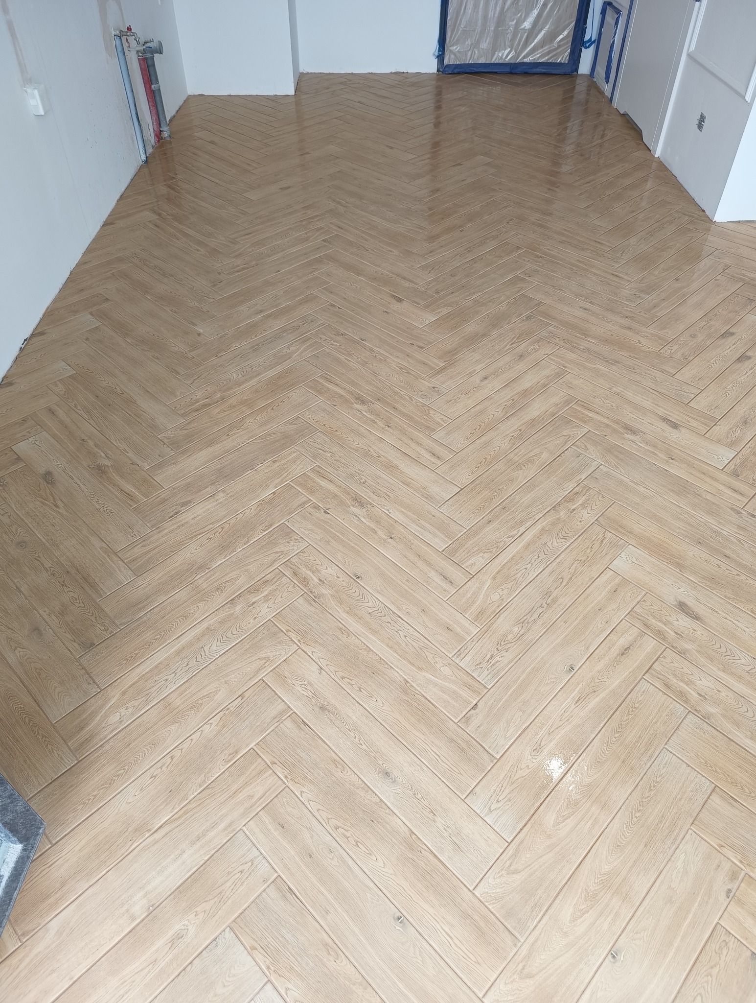 Ułożona w jodełkę, jasna, drewnopodobna podłoga z płytek ceramicznych w trakcie prac wykończeniowych w łazience, widoczne rury instalacji wodnej przy ścianie.