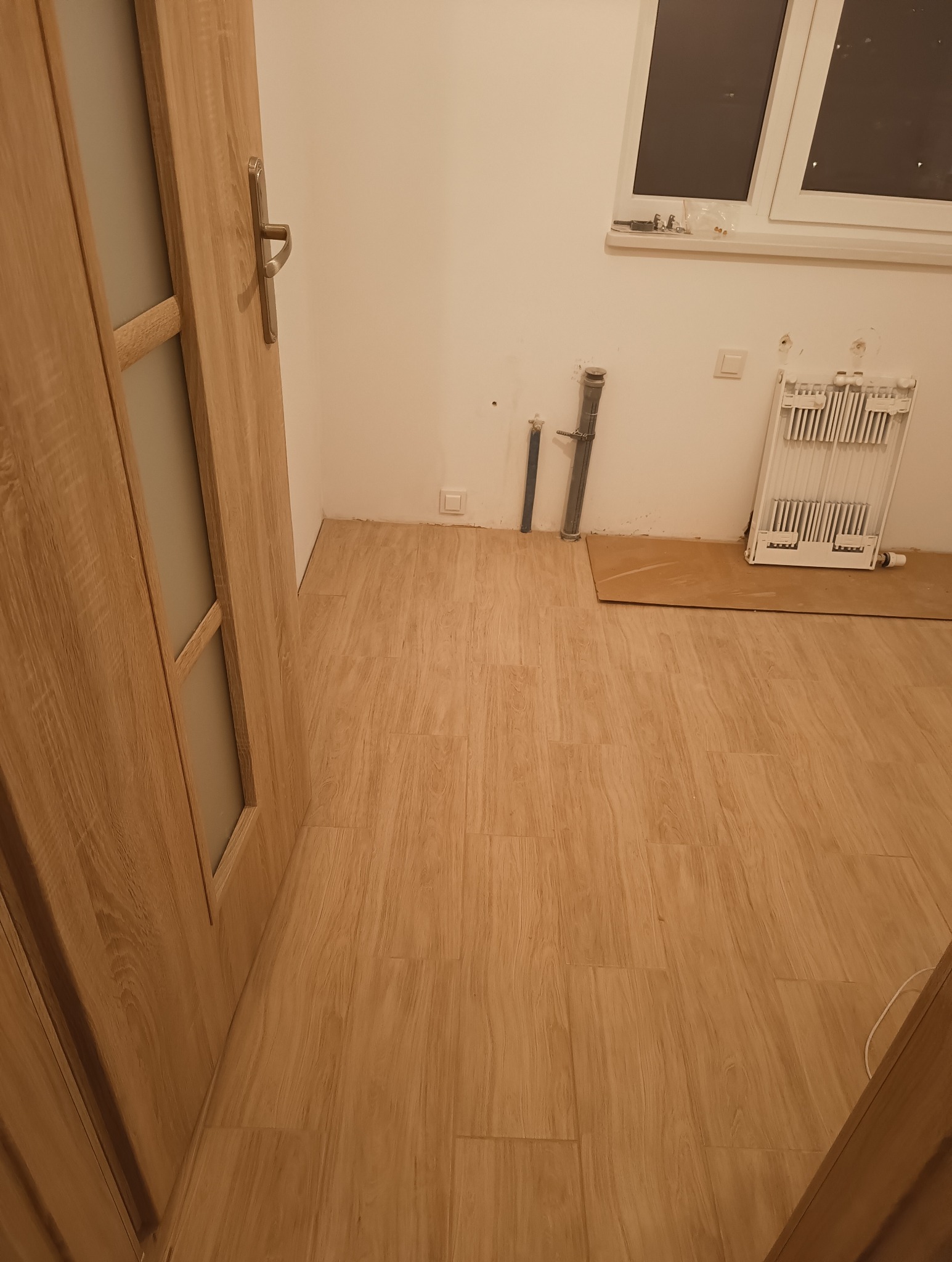 Niedokończona łazienka z nową, drewnopodobną podłogą, grzejnikiem i rurami wystającymi ze ściany, widoczne przygotowania do dalszych prac.