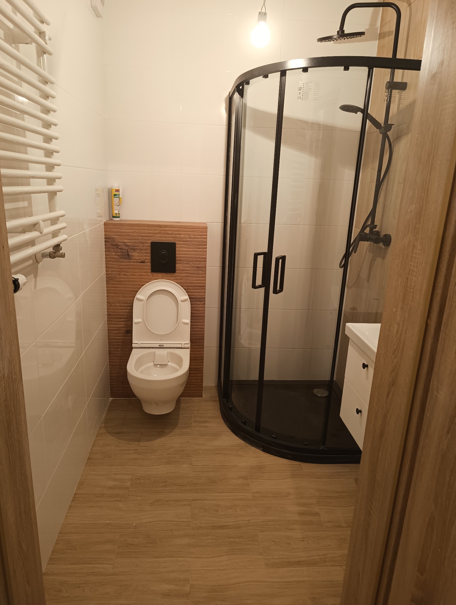 Nowoczesna łazienka z kabiną prysznicową typu walk-in z czarnymi profilami, podwieszaną toaletą z drewnianym panelem dekoracyjnym, grzejnikiem drabinkowym i drewnopodobną podłogą.