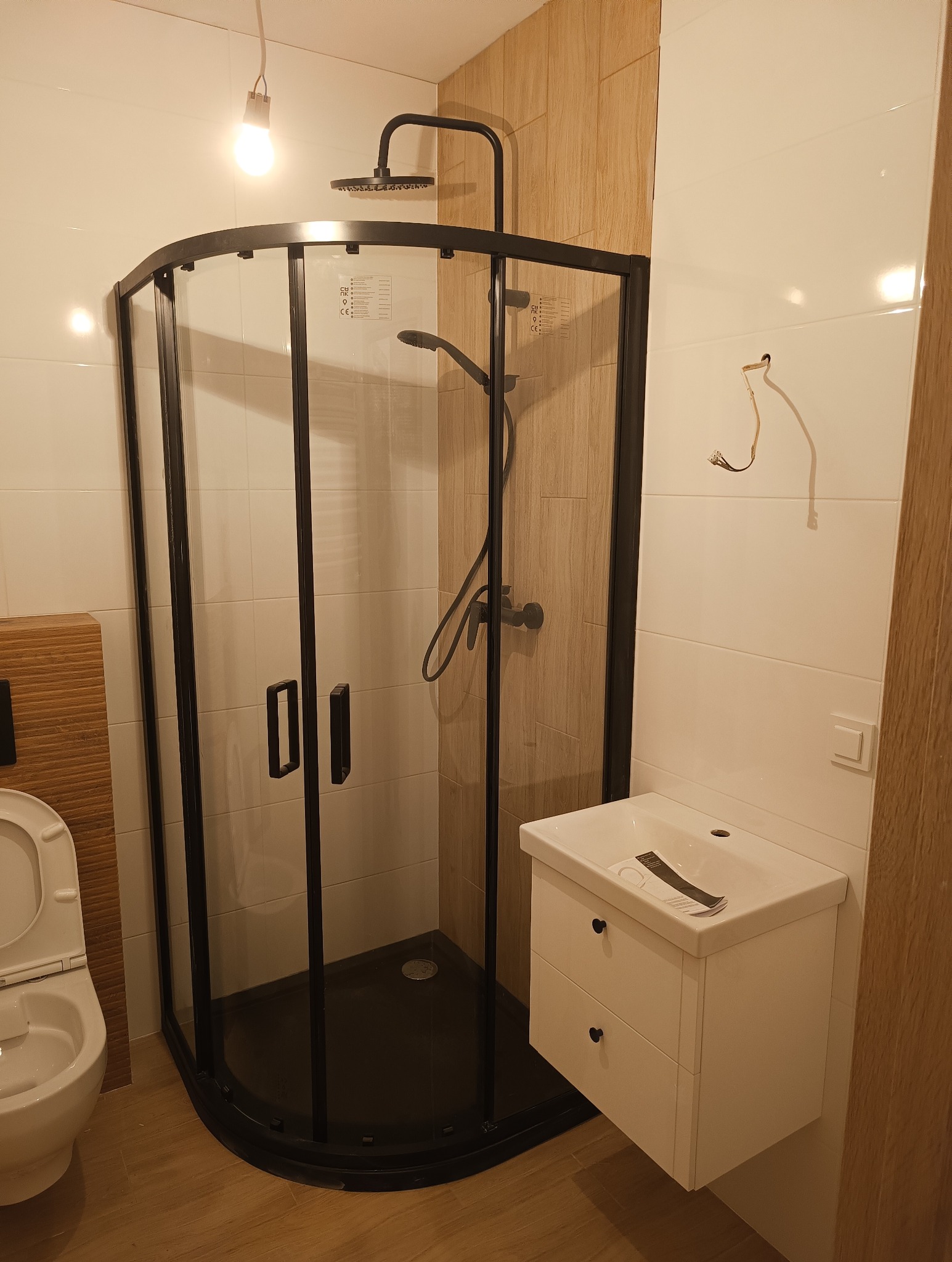 Nowoczesna kabina prysznicowa z czarną ramą, biała umywalka z szafką, wisząca żarówka i częściowo widoczna toaleta w wykończonej łazience z jasnymi płytkami.