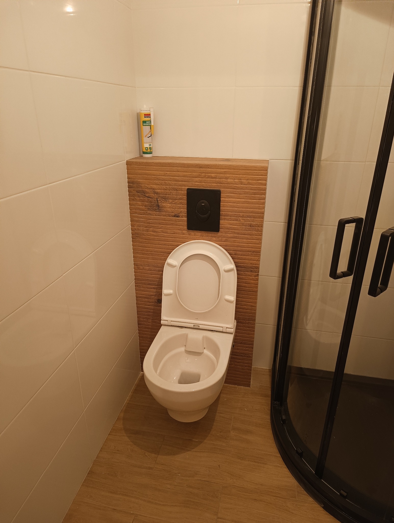 Wisząca muszla klozetowa z otwartą deską, zamontowana na drewnopodobnej płycie z czarnym przyciskiem spłukującym, obok kabiny prysznicowej z czarną ramą i przezroczystym szkłem, na podłodze...