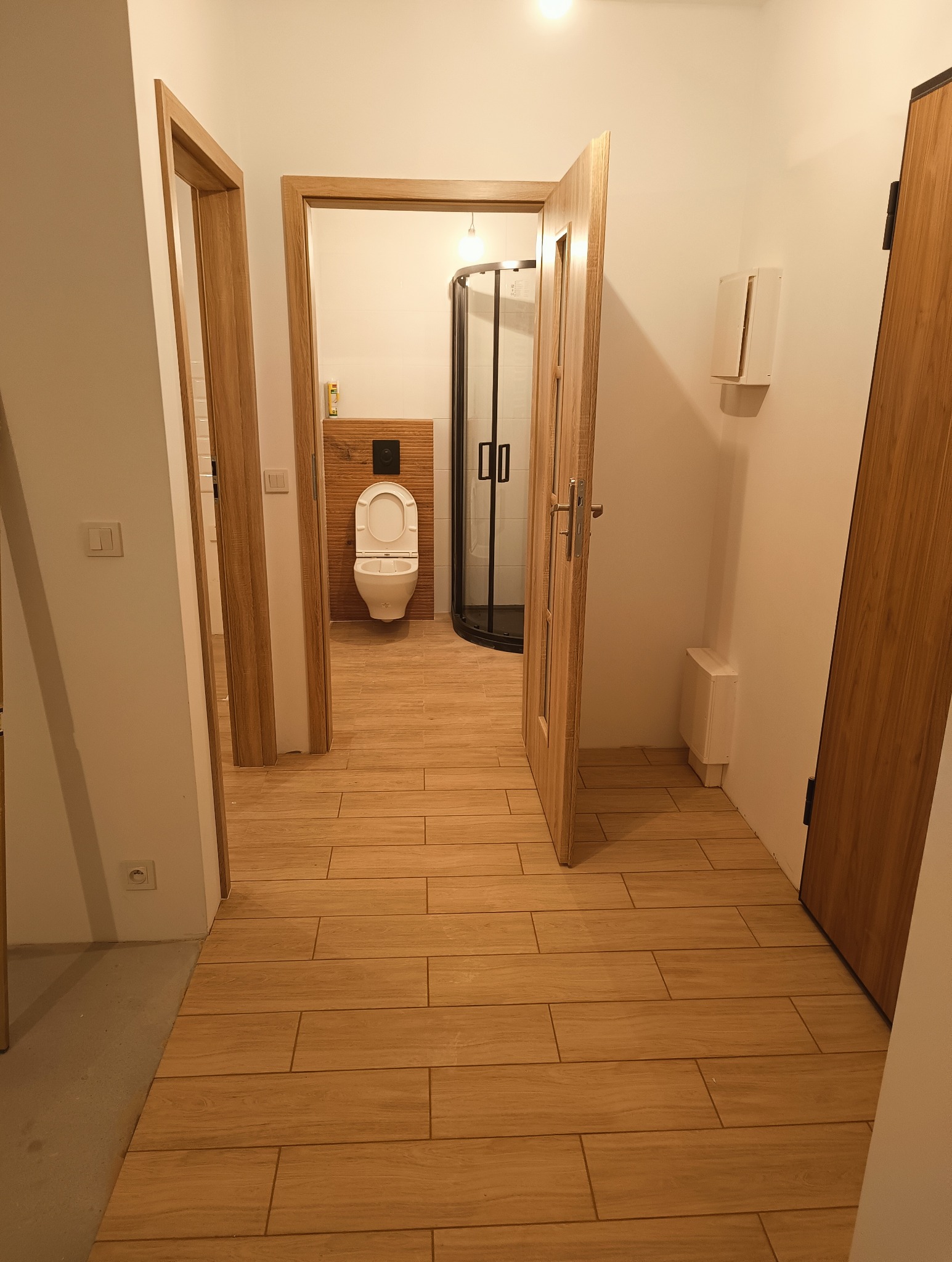 Widok przez otwarte drzwi na wykończoną łazienkę z kabiną prysznicową i toaletą, wykończoną płytkami imitującymi drewno na podłodze i ścianie za toaletą.