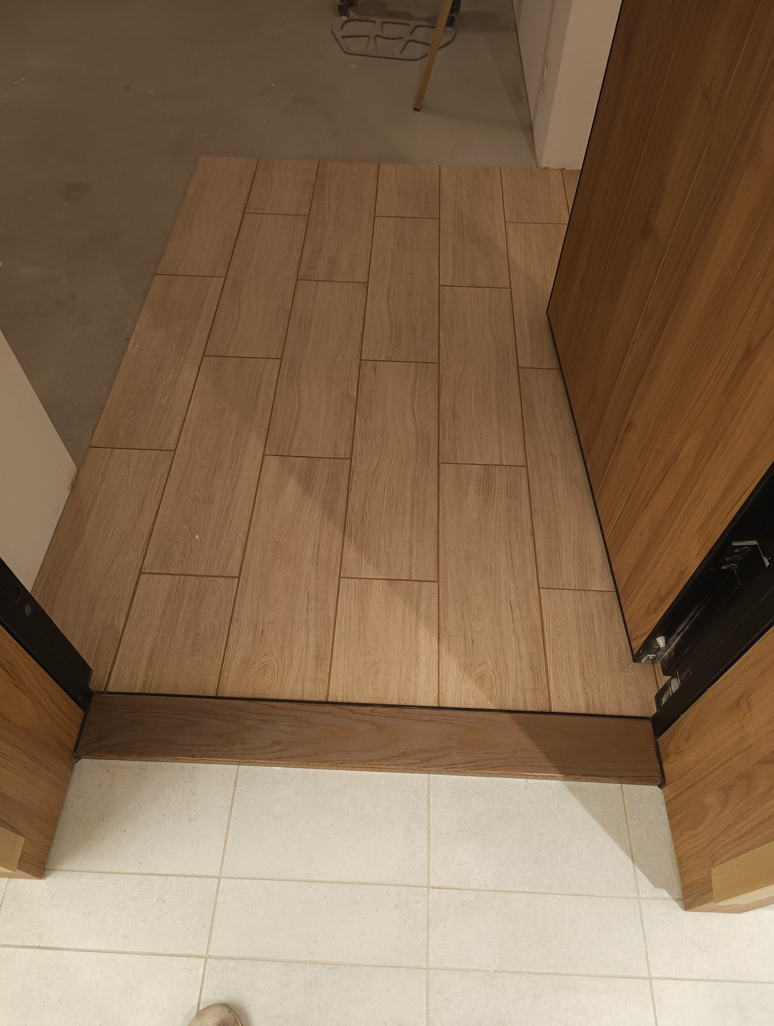 Ułożona podłoga z płytek imitujących drewno w łazience, widok z góry, listwa przypodłogowa z ciemnego drewna, w tle białe płytki podłogowe i fragment drzwi.