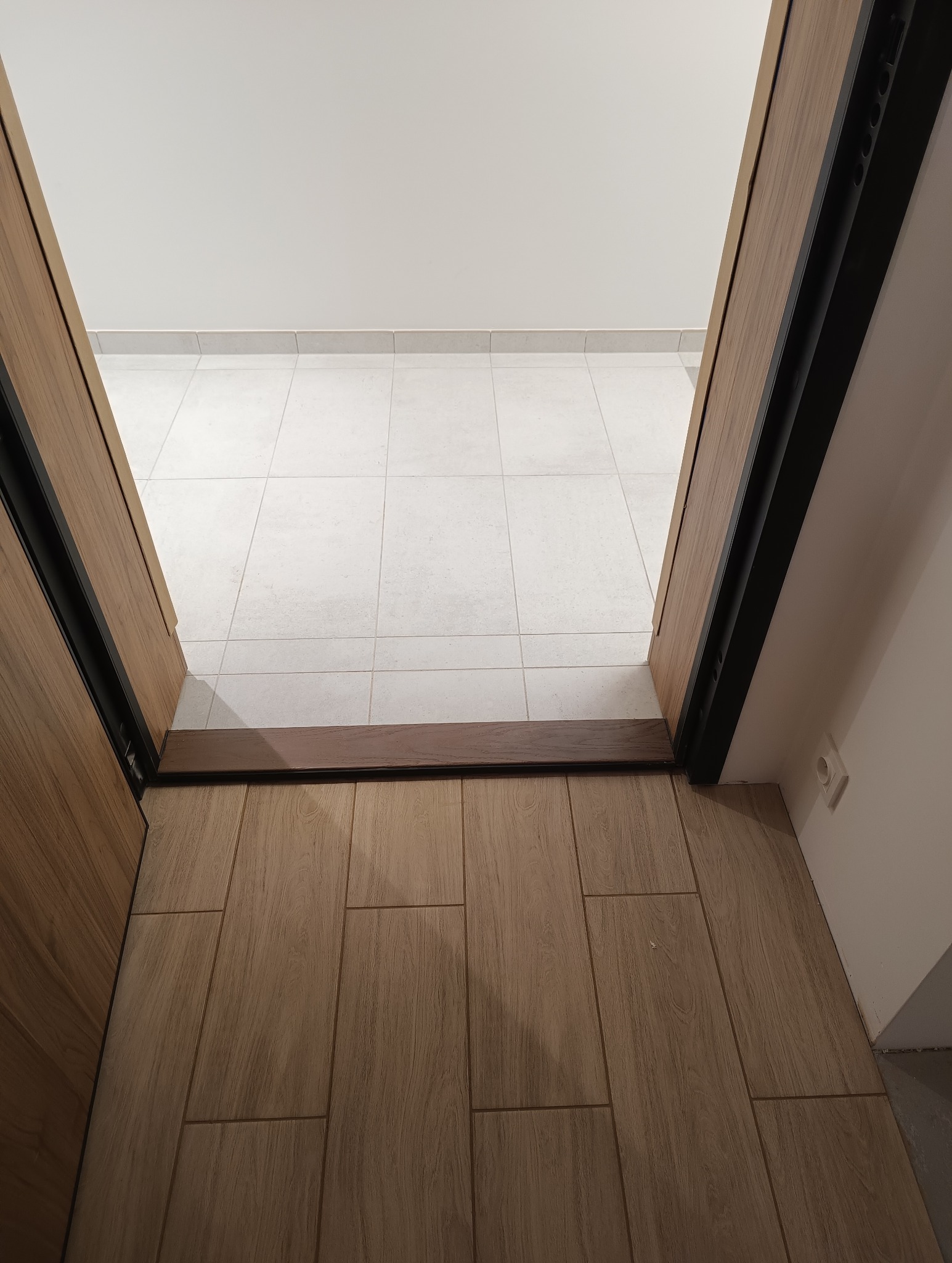 Widok z góry na próg drzwiowy oddzielający dwie różne powierzchnie podłogowe: jasne, kwadratowe płytki w oddali i podłużne, drewnopodobne płytki na pierwszym planie. Drzwi z drewnianą ramą...