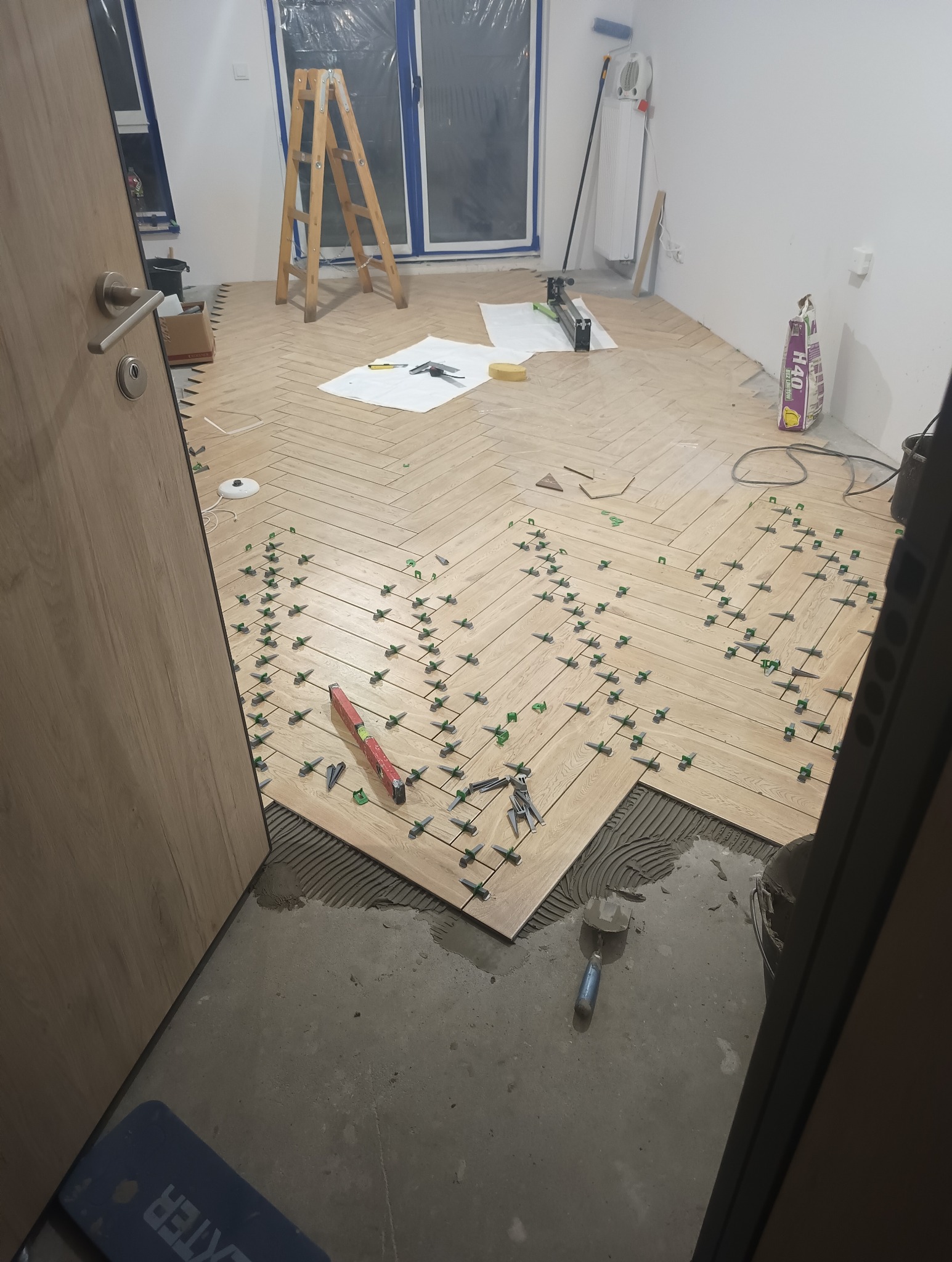 Układanie parkietu z płytek podłogowych w jodełkę, widoczne zielone krzyżyki dystansowe, poziomica, szpachelka, oraz narzędzia na białym materiale zabezpieczającym.