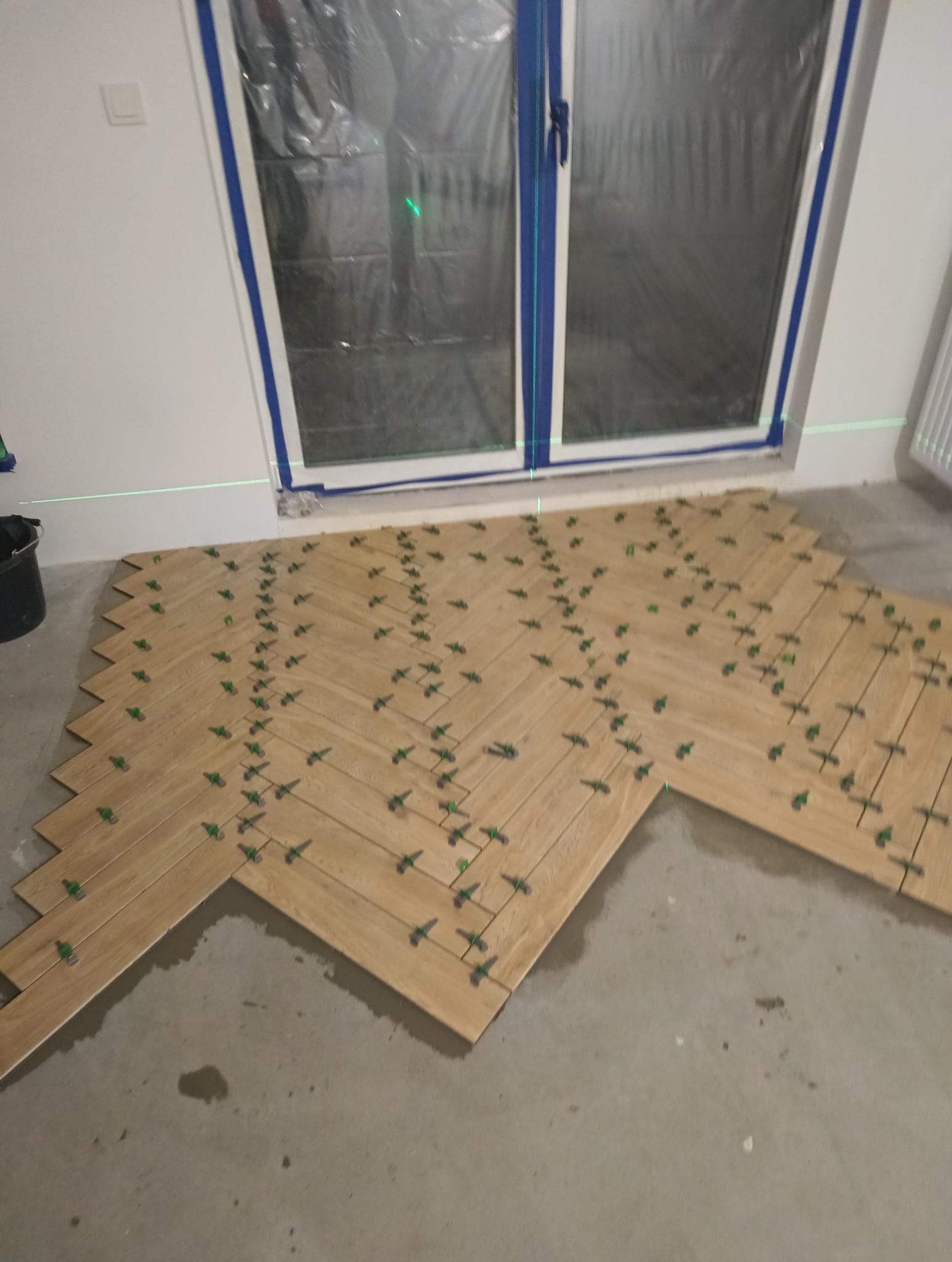 Układanie jasnych, drewnopodobnych płytek w jodełkę w łazience, widoczne zielone krzyżyki dystansowe, okno zabezpieczone folią malarską, poziomica laserowa.