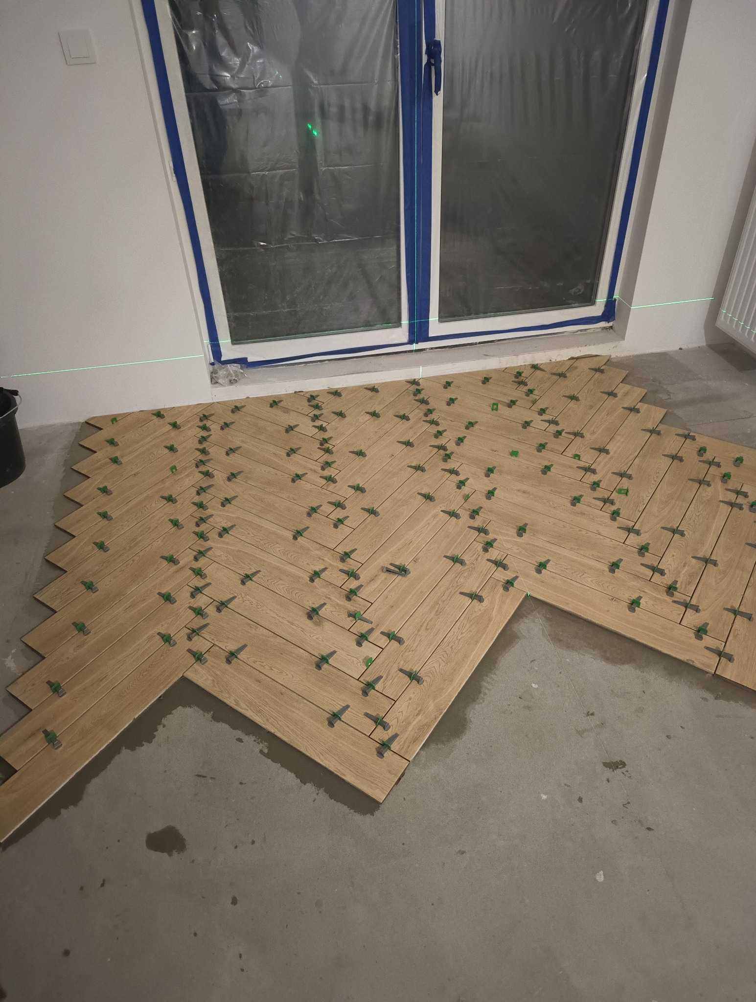 Fragment podłogi w trakcie układania płytek imitujących drewno w jodełkę, widoczne zielone krzyżyki dystansowe i poziomica laserowa, okno zabezpieczone folią.