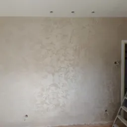 Ściana po nałożeniu dekoracyjnej, beżowej powłoki z efektem subtelnych przetarć, widoczne kable wystające z sufitu, zakryte folią drzwi i aluminiowa drabina.
