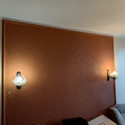 Ściana w kolorze terakoty z wyraźną fakturą, ozdobiona ramą i dwoma kinkietami w kształcie kwiatów, oświetlającymi fragment pokoju.