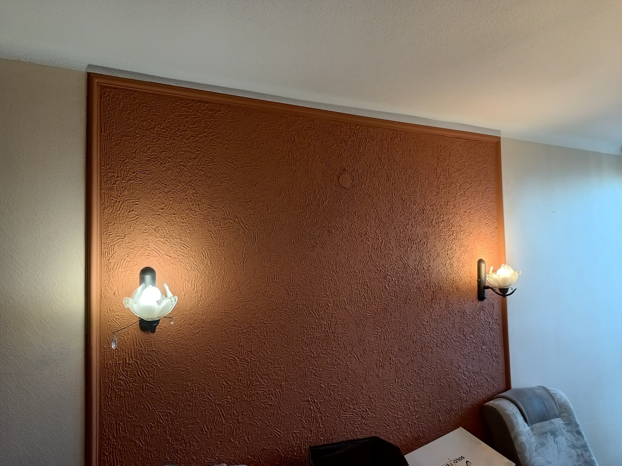 Ściana w kolorze terakoty z wyraźną fakturą, ozdobiona ramą i dwoma kinkietami w kształcie kwiatów, oświetlającymi fragment pokoju.