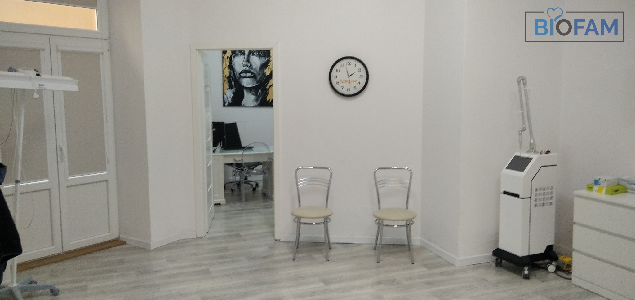 Jasne wnętrze poczekalni kliniki z dwoma krzesłami, zegarem ściennym, obrazem i urządzeniem kosmetycznym na kółkach, widoczny gabinet w tle.