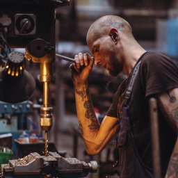 Fomin-Art - Tatuowany mężczyzna w kombinezonie obsługuje wiertarkę kolumnową, skupiony na precyzyjnym wierceniu metalowego elementu w warsztacie.