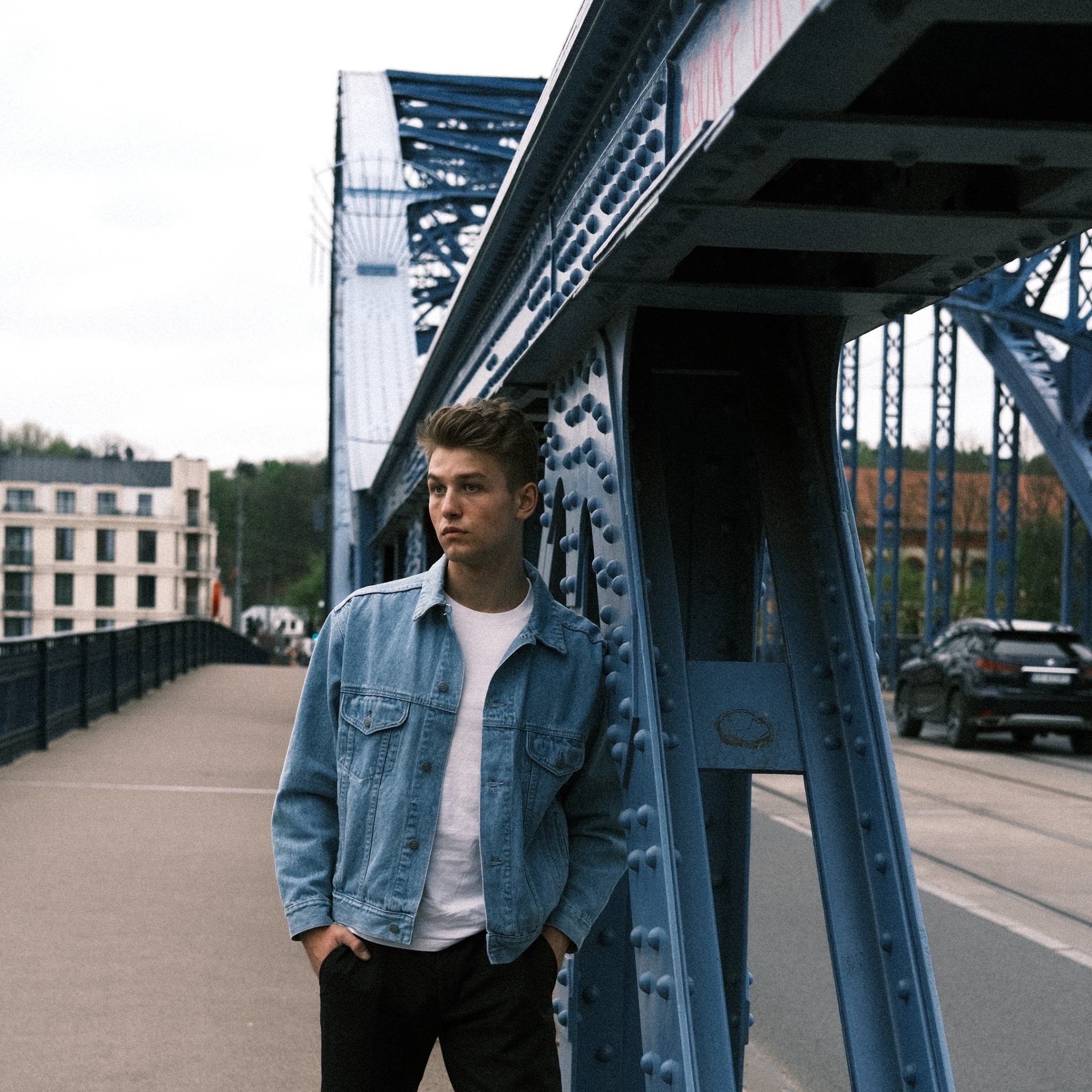 Młody mężczyzna w jeansowej kurtce opiera się o błękitną, nitowaną konstrukcję mostu, z widokiem na budynki i przejeżdżający samochód w tle.