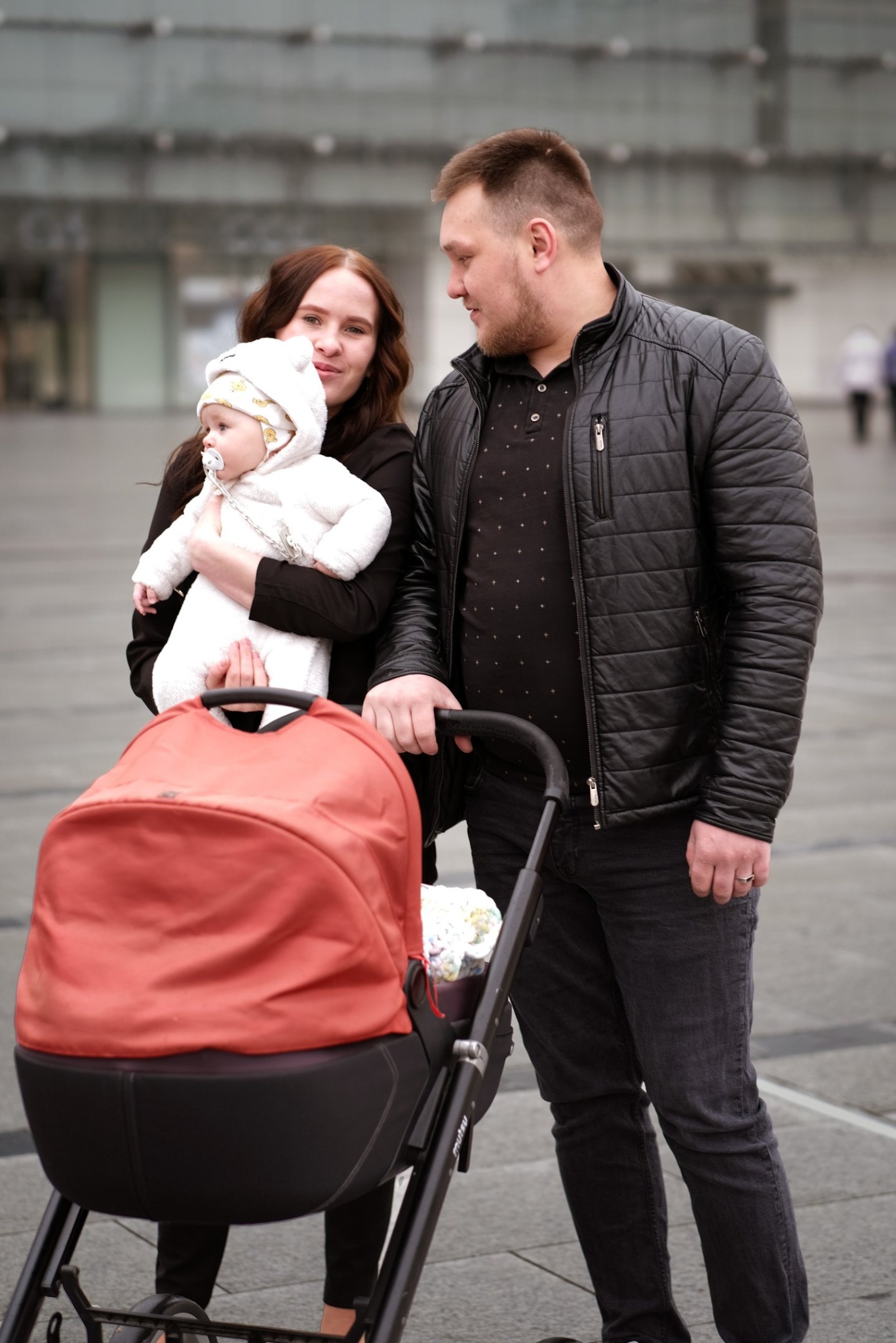 Uśmiechnięta rodzina z małym dzieckiem w białym kombinezonie i wózkiem na tle nowoczesnego budynku.