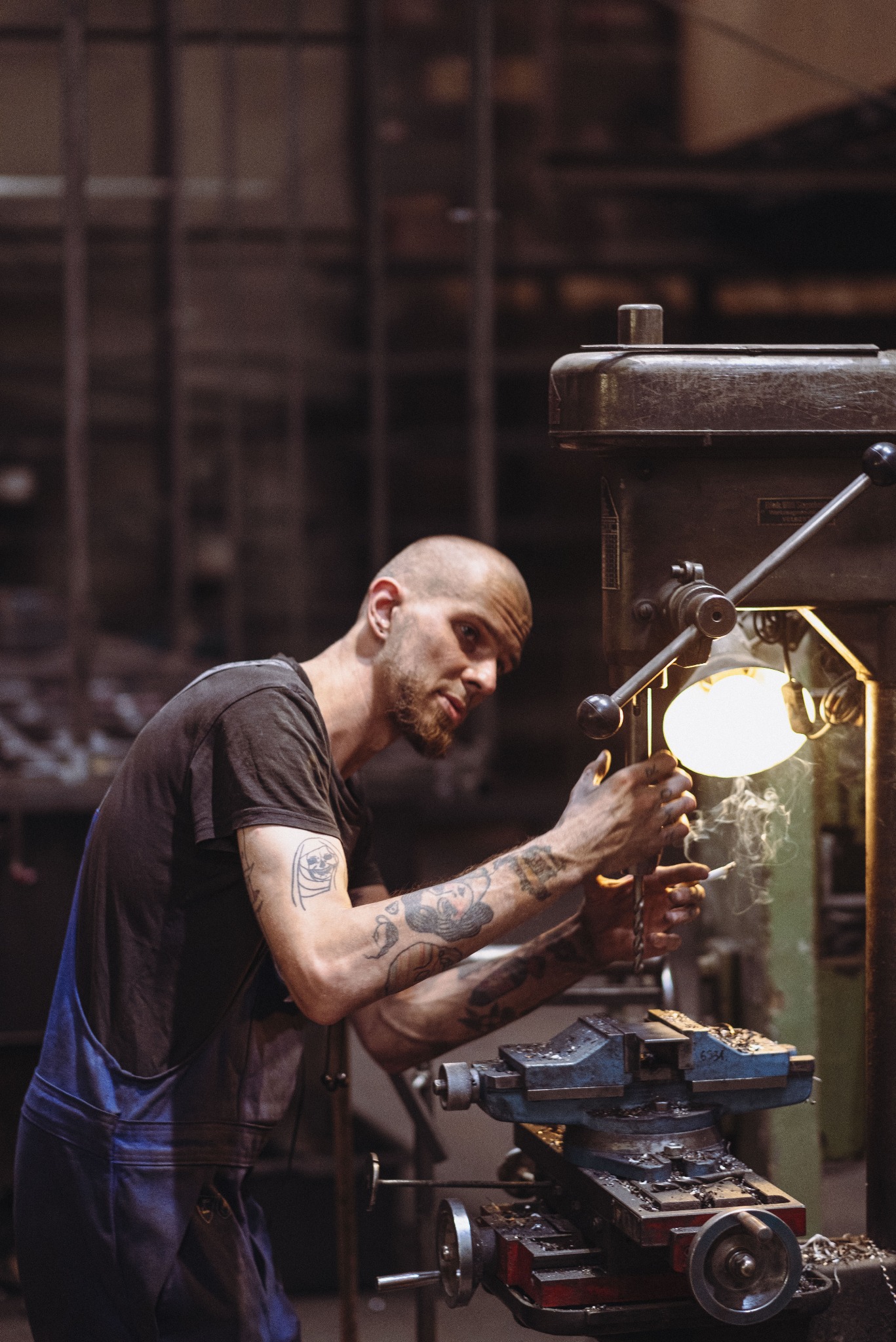Pracownik z tatuażami obsługuje wiertarkę stołową, w tle widoczne metalowe konstrukcje.