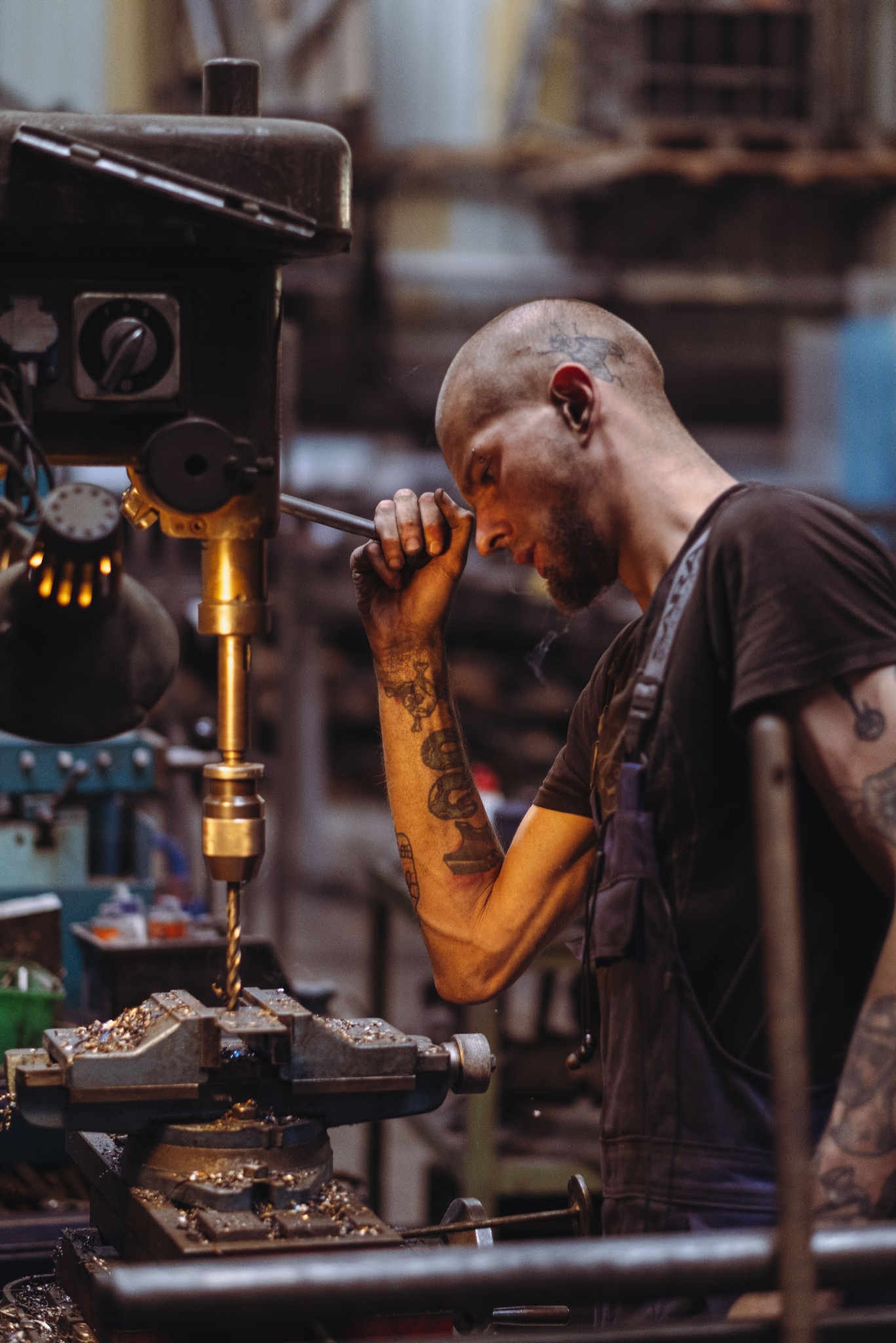 Tatuowany mężczyzna w kombinezonie obsługuje wiertarkę kolumnową, skupiony na precyzyjnym wierceniu metalowego elementu w warsztacie.
