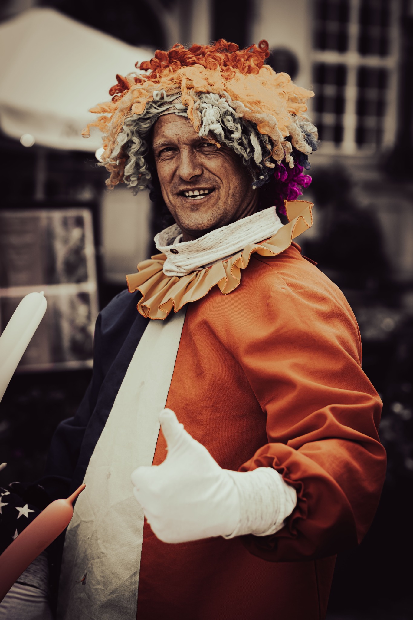 Uśmiechnięty mężczyzna w kostiumie klauna z kolorową peruką i balonami modelowanymi, kciuk w górę.