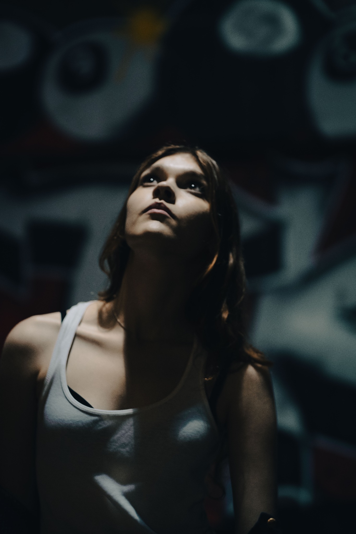 Portret kobiety spoglądającej w górę, w tle rozmyte graffiti na ścianie, oświetlenie od dołu.