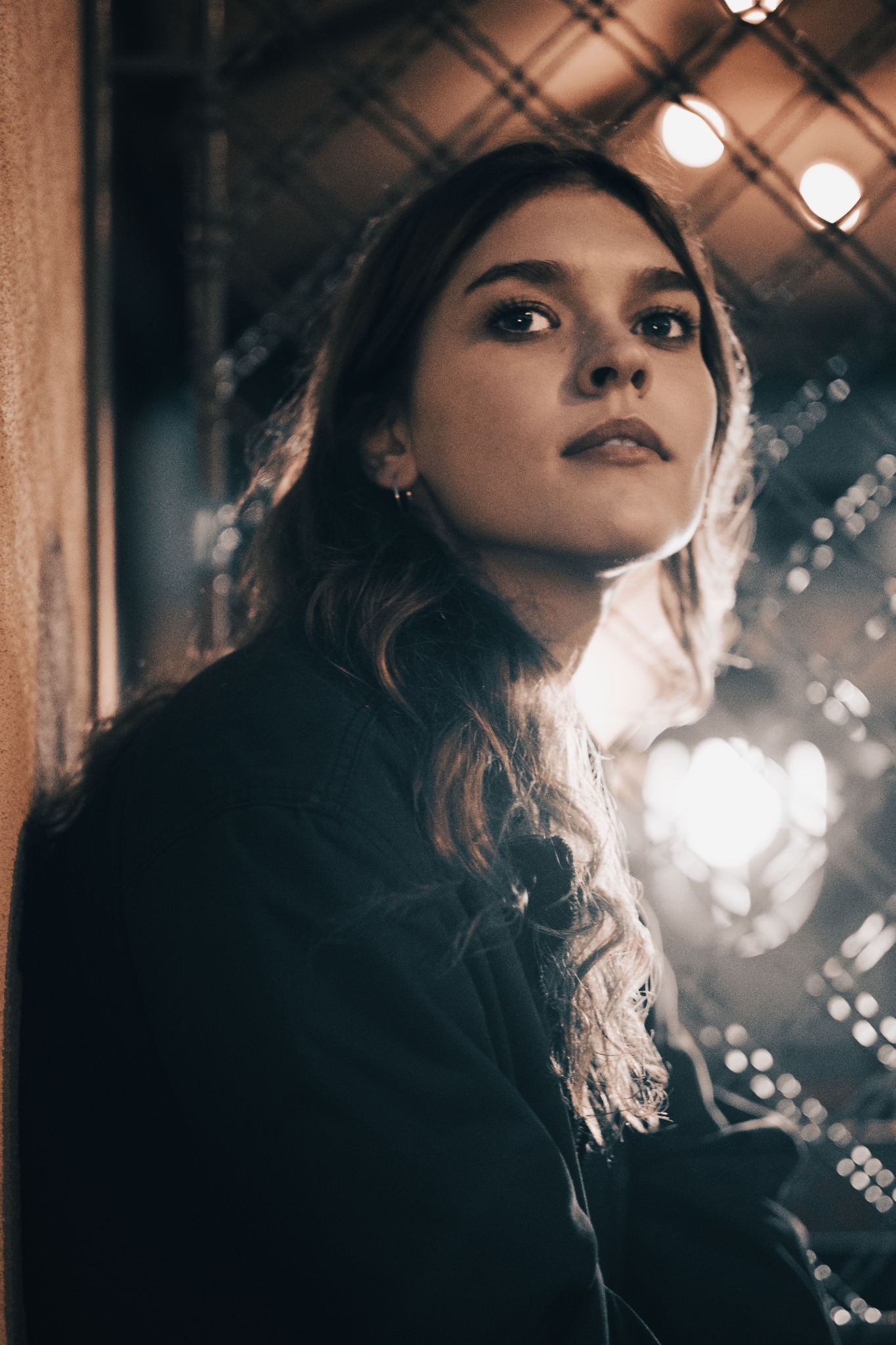 Portret młodej kobiety z delikatnym makijażem, patrzącej w górę, z widoczną kolczykiem w uchu, na tle rozmytych świateł i tekstury ściany.