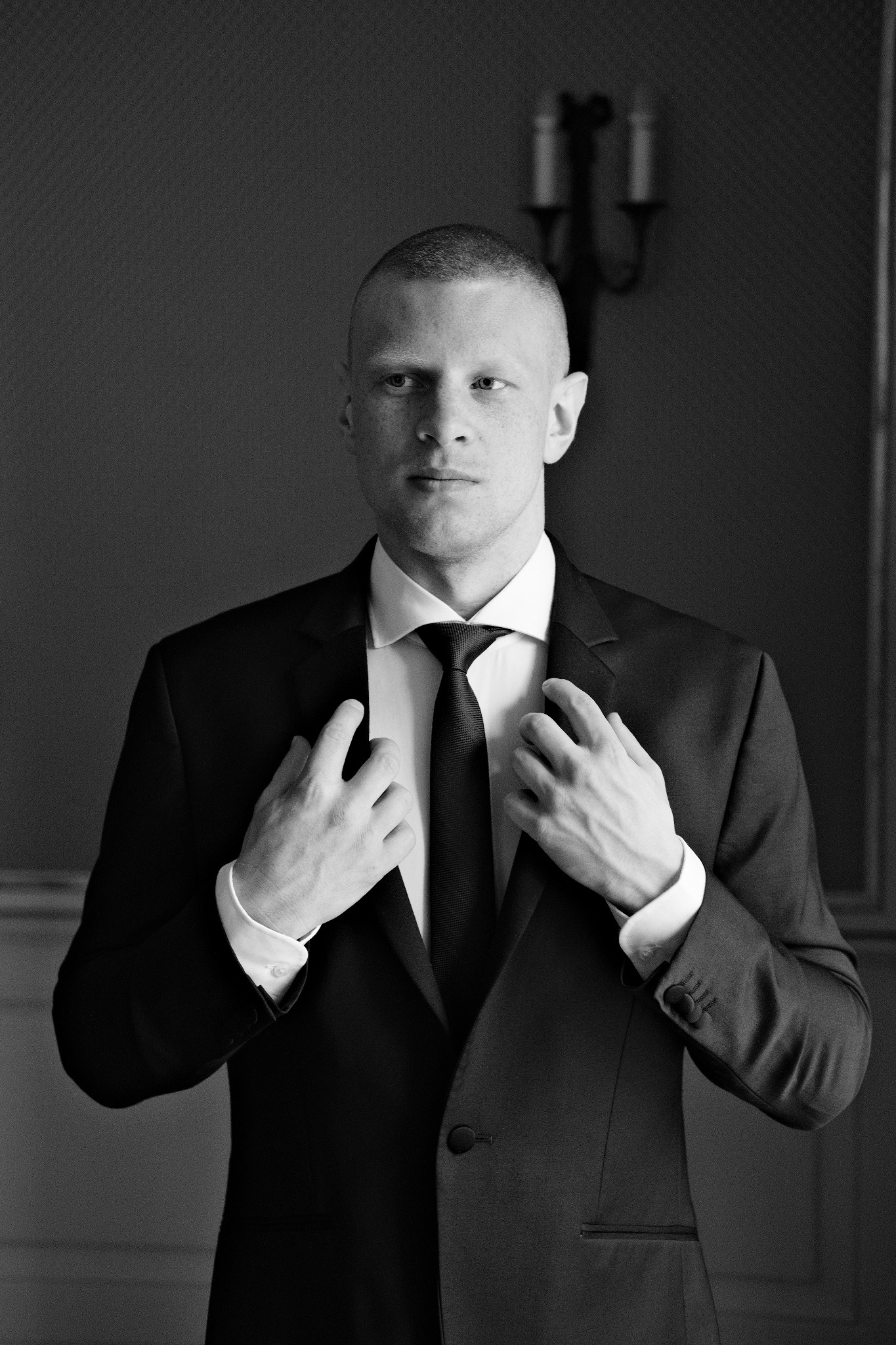 Mężczyzna w czarno-białym portrecie, poprawiający marynarkę i krawat, z widocznym świecznikiem w tle.