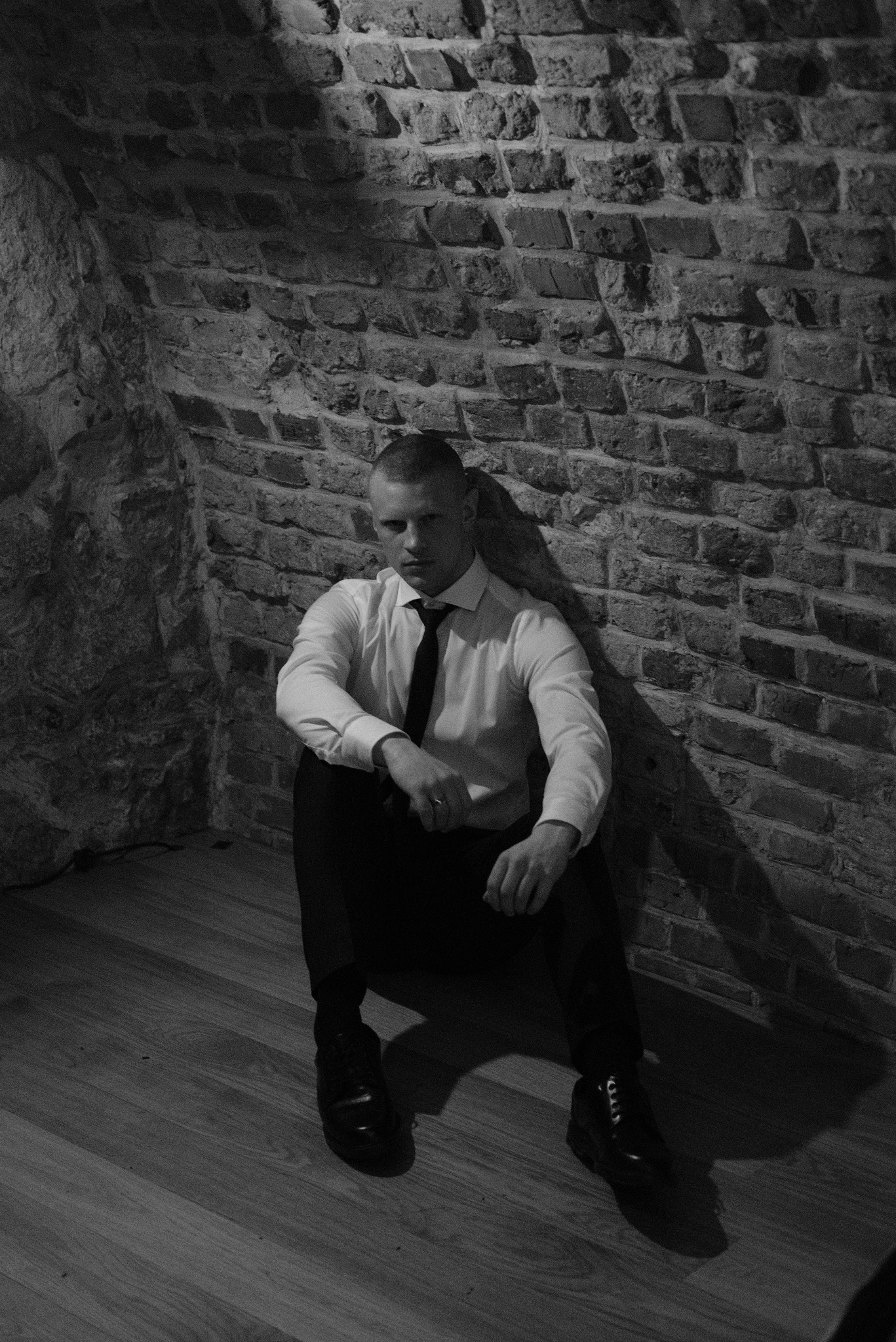 Mężczyzna w białej koszuli i krawacie siedzi na podłodze oparty o ceglaną ścianę w zacienionym wnętrzu.