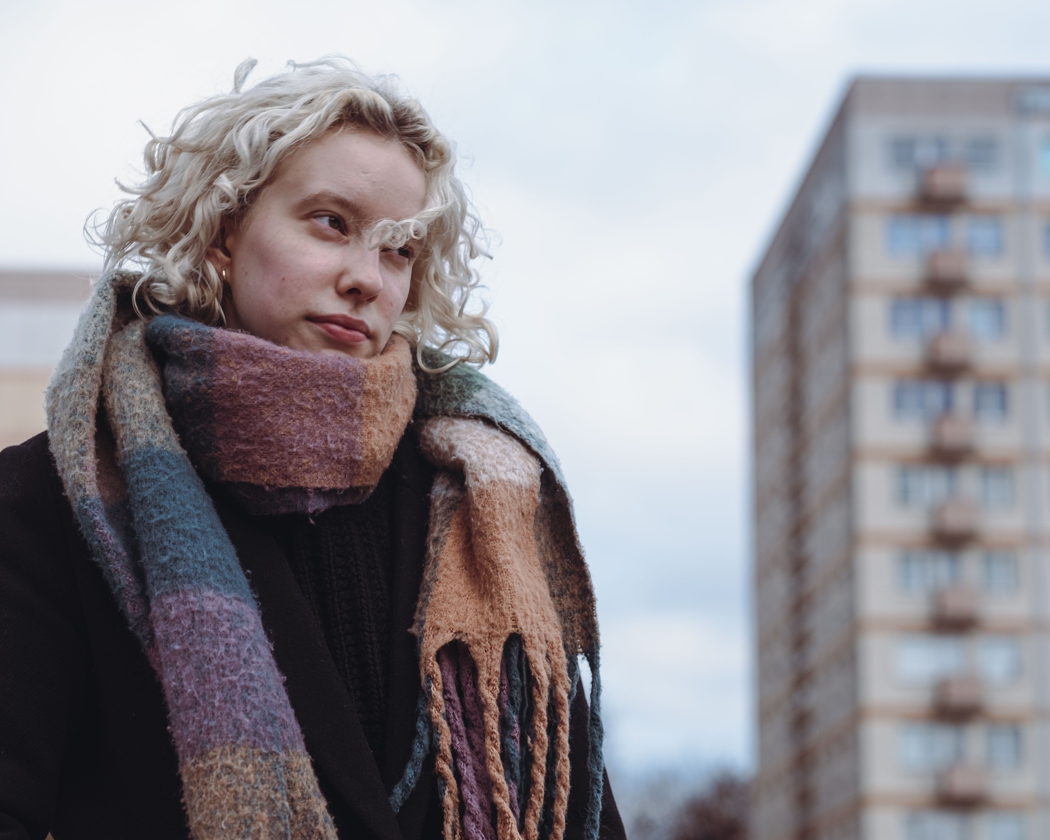 Portret kobiety z kręconymi blond włosami, w grubym, kolorowym szaliku na tle bloku mieszkalnego.