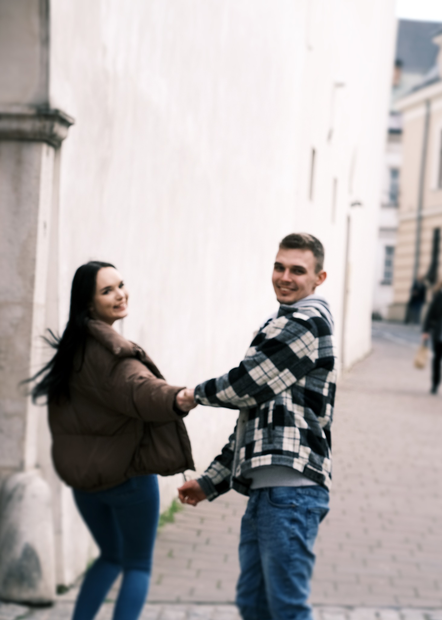 Uśmiechnięta para trzymająca się za ręce, odwrócona w stronę fotografa na tle miejskiej uliczki z jasną ścianą.