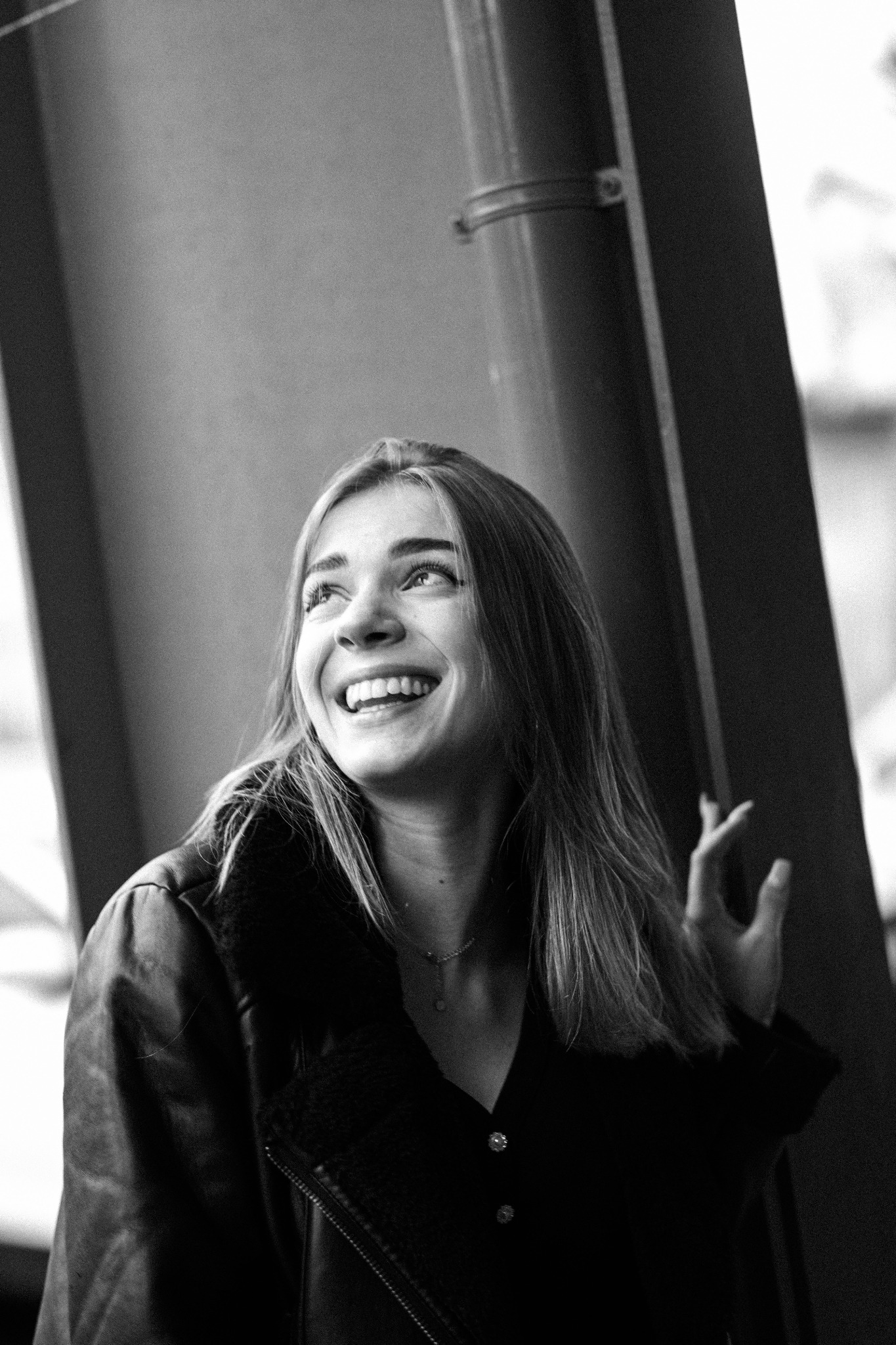 Portret uśmiechniętej młodej kobiety w czarno-białej tonacji, spoglądającej w górę, ubrana w skórzaną kurtkę z futrzanym kołnierzem, sfotografowana z bliska na tle ściany i metalowej konstrukcji.